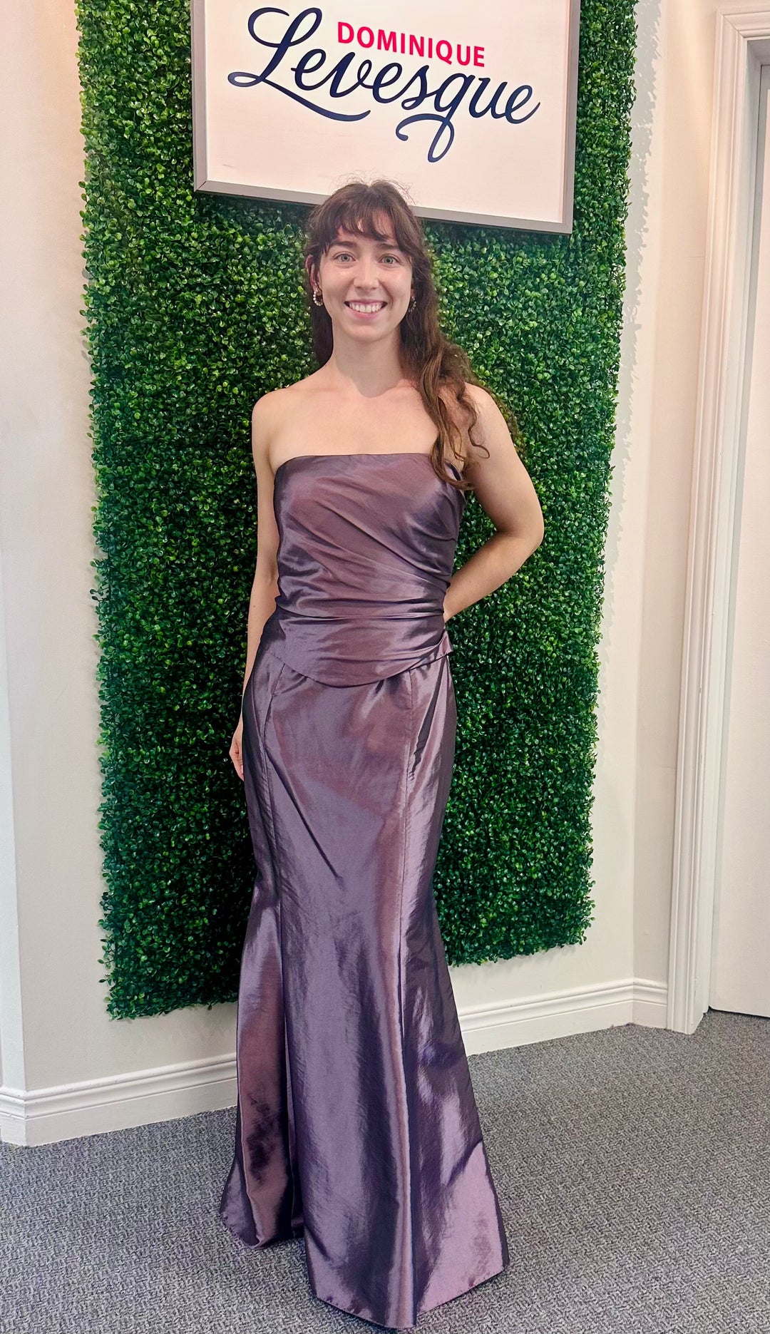 Woman in a plum  strapless gown standing in front of a green wall with a sign above.