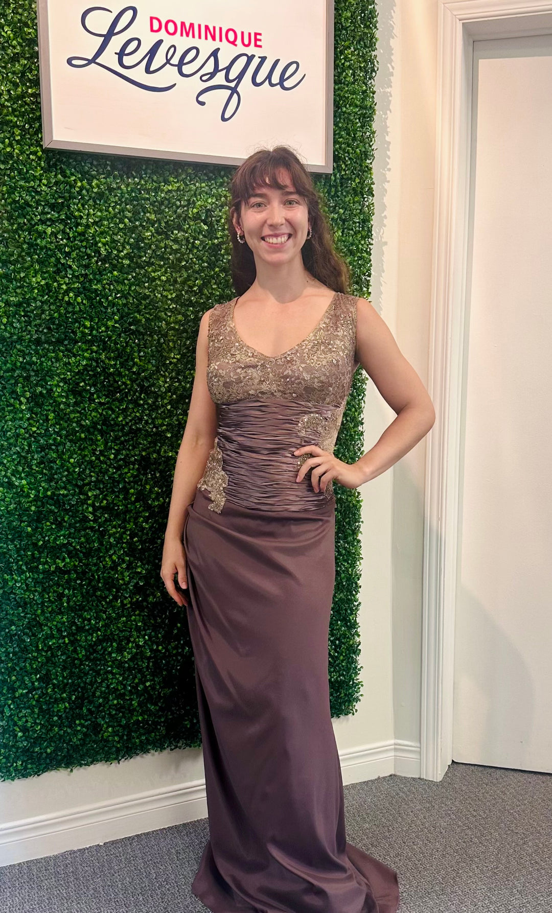 Woman in a formal bronze gown standing in front of a green wall with a sign above.