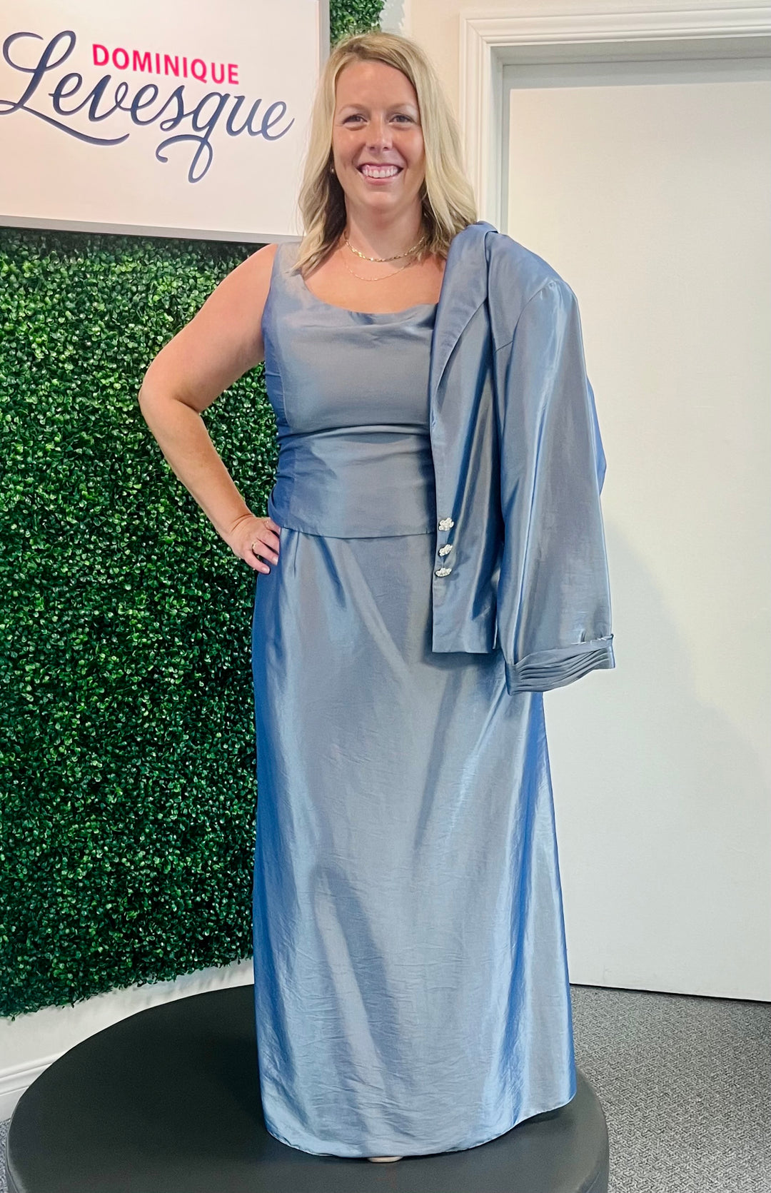 Woman in a light blue dress with a matching jacket standing in front of a green wall with 'Dominique Levesque' branding.