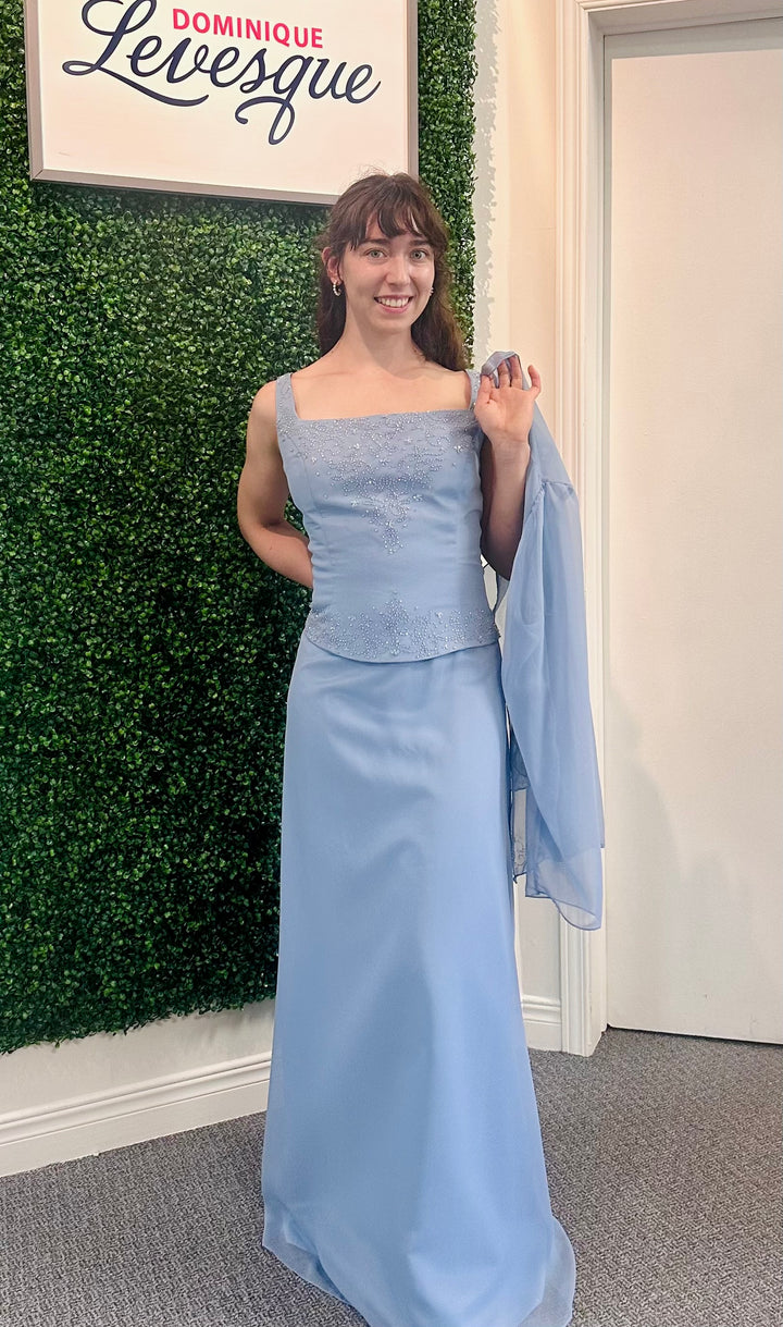 Woman in a light blue evening gown holding a matching vest against a green wall with a sign saying &