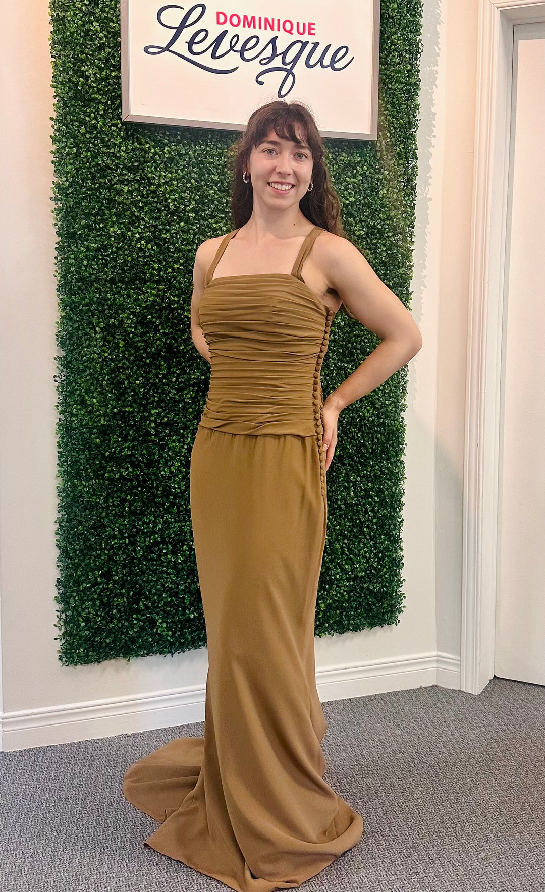 Woman in a mustard brown dress standing in front of a green wall with a sign above it saying "Dominque Levesque".