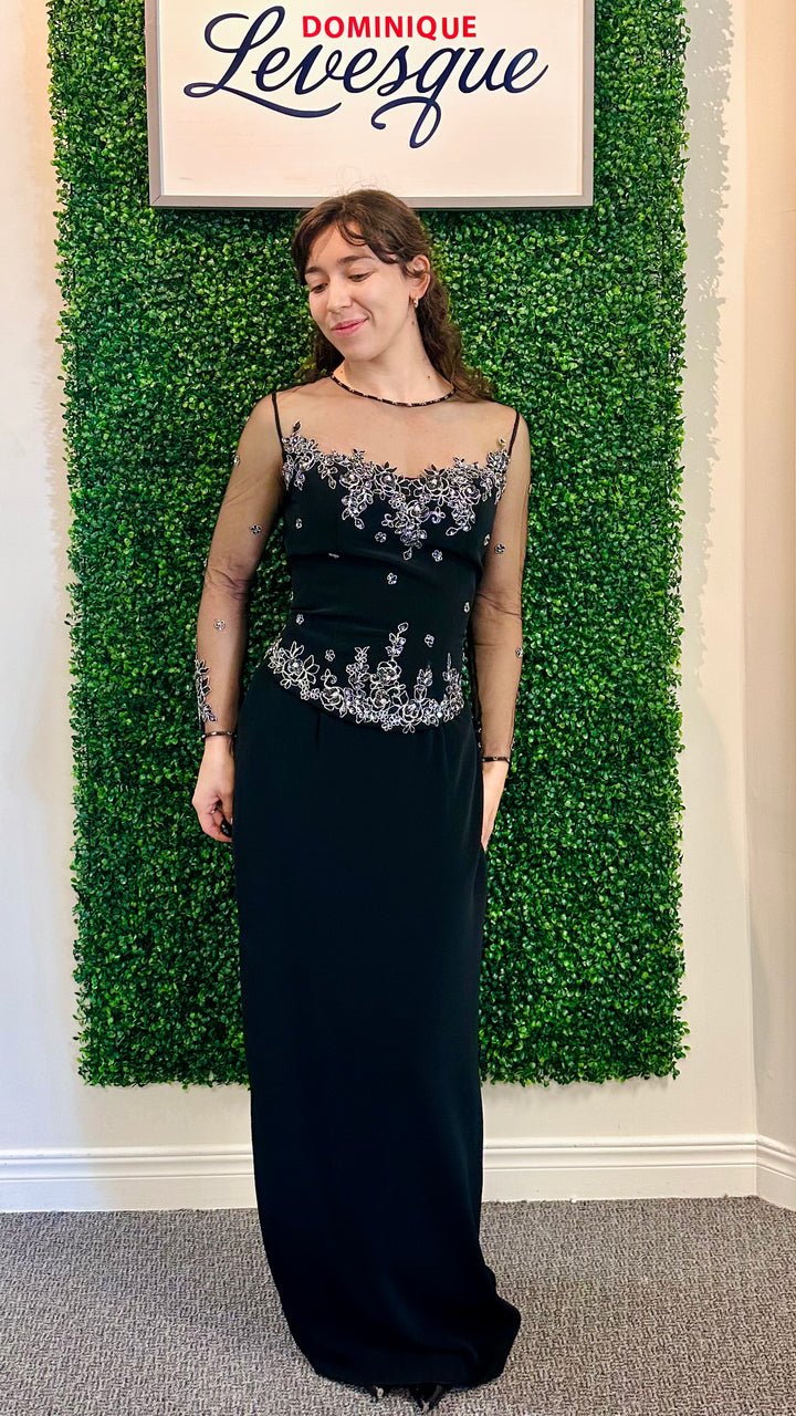 Woman in a black evening gown with silver embellishments standing in front of a green leafy wall with &