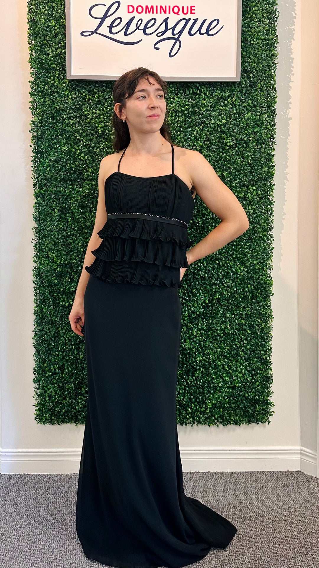 Woman in a black dress standing against a green leafy wall with a sign above.