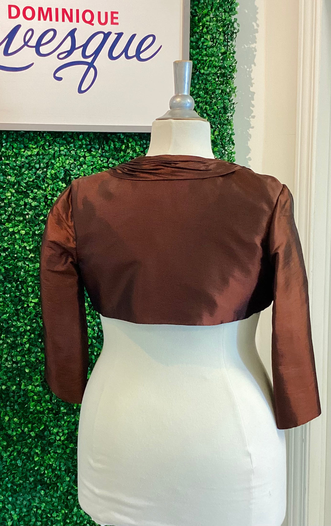 Brown satin bolero jacket on a mannequin against a green leafy background with &