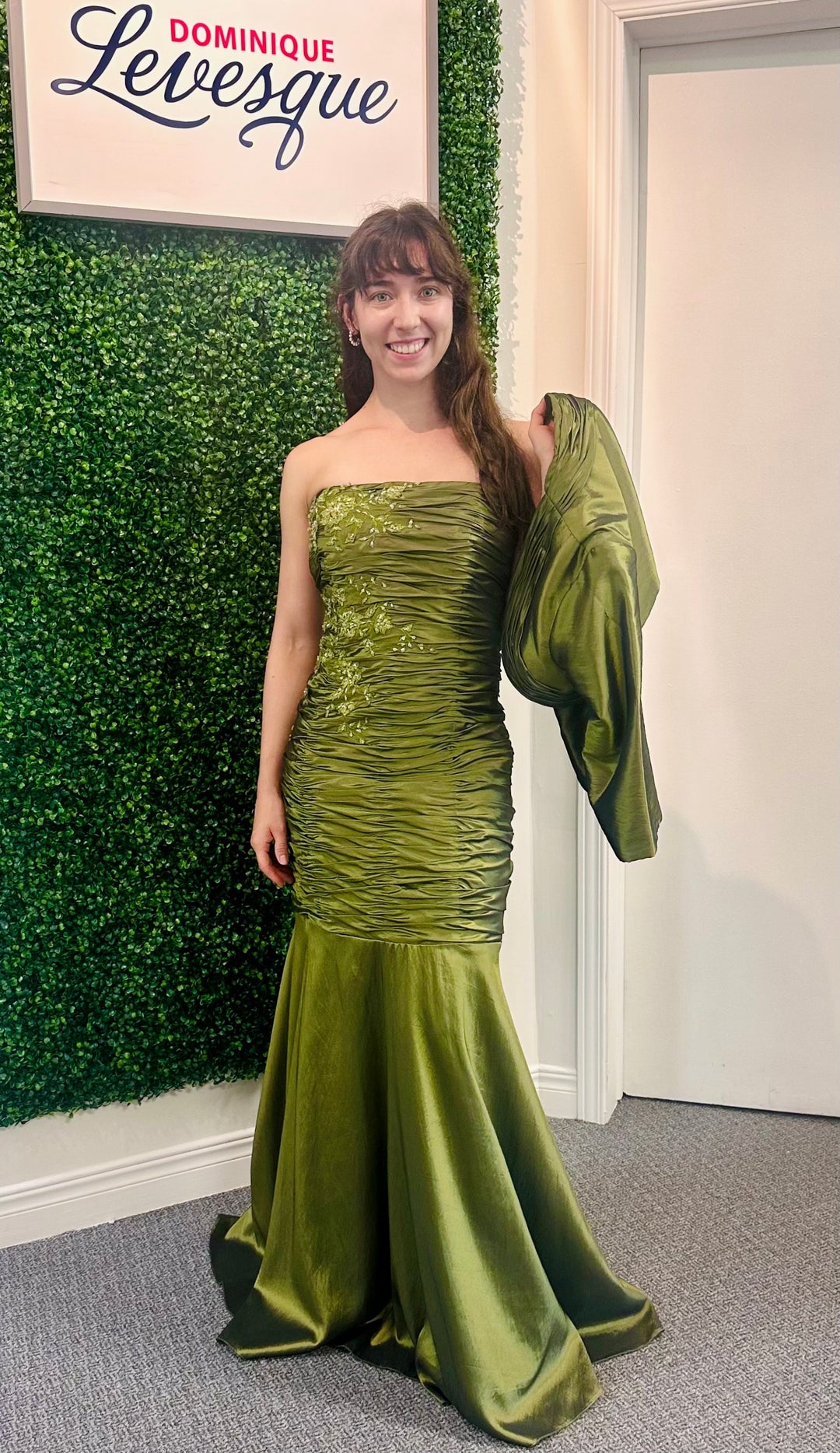 Woman in a green evening gown standing against a green wall with 'Dominique Levesque' branding.