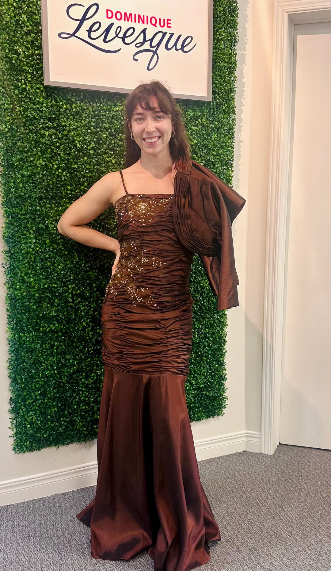 Woman in a brown evening gown standing in front of a green wall with a sign above.