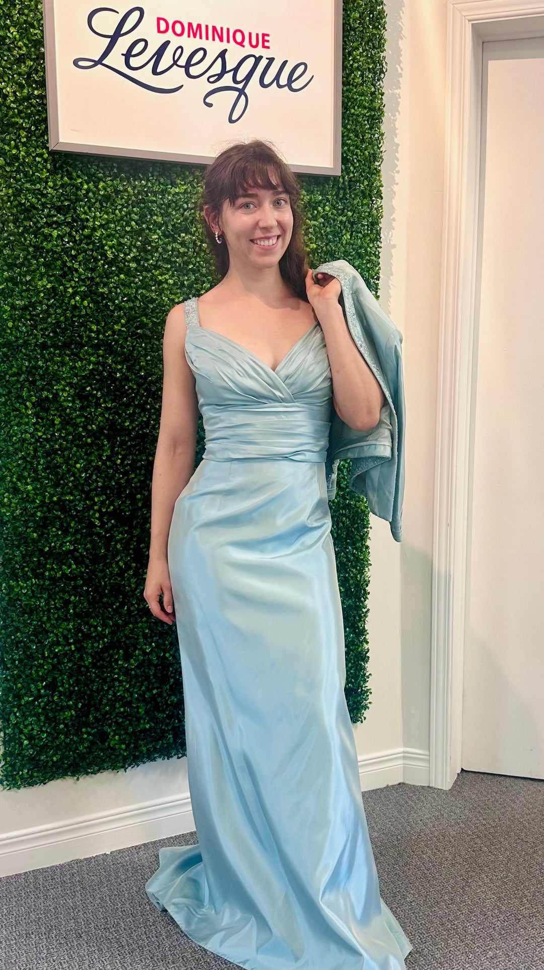 Woman in a light blue evening gown standing against a green leafy wall with a sign above saying 'Dominique Levesque'.