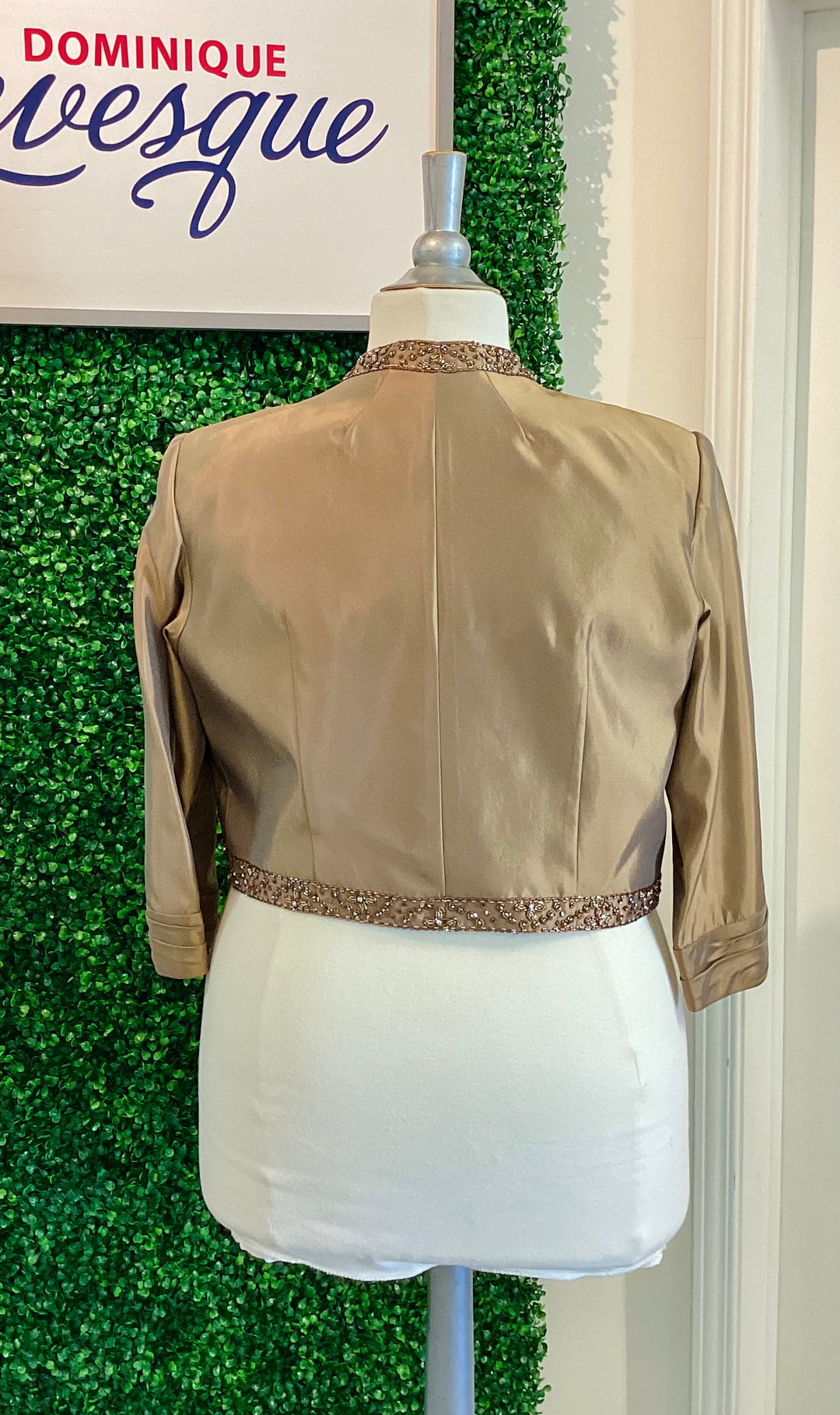 Mannequin wearing a bronze vest with decorative trim against a green leafy background, featuring the brand &