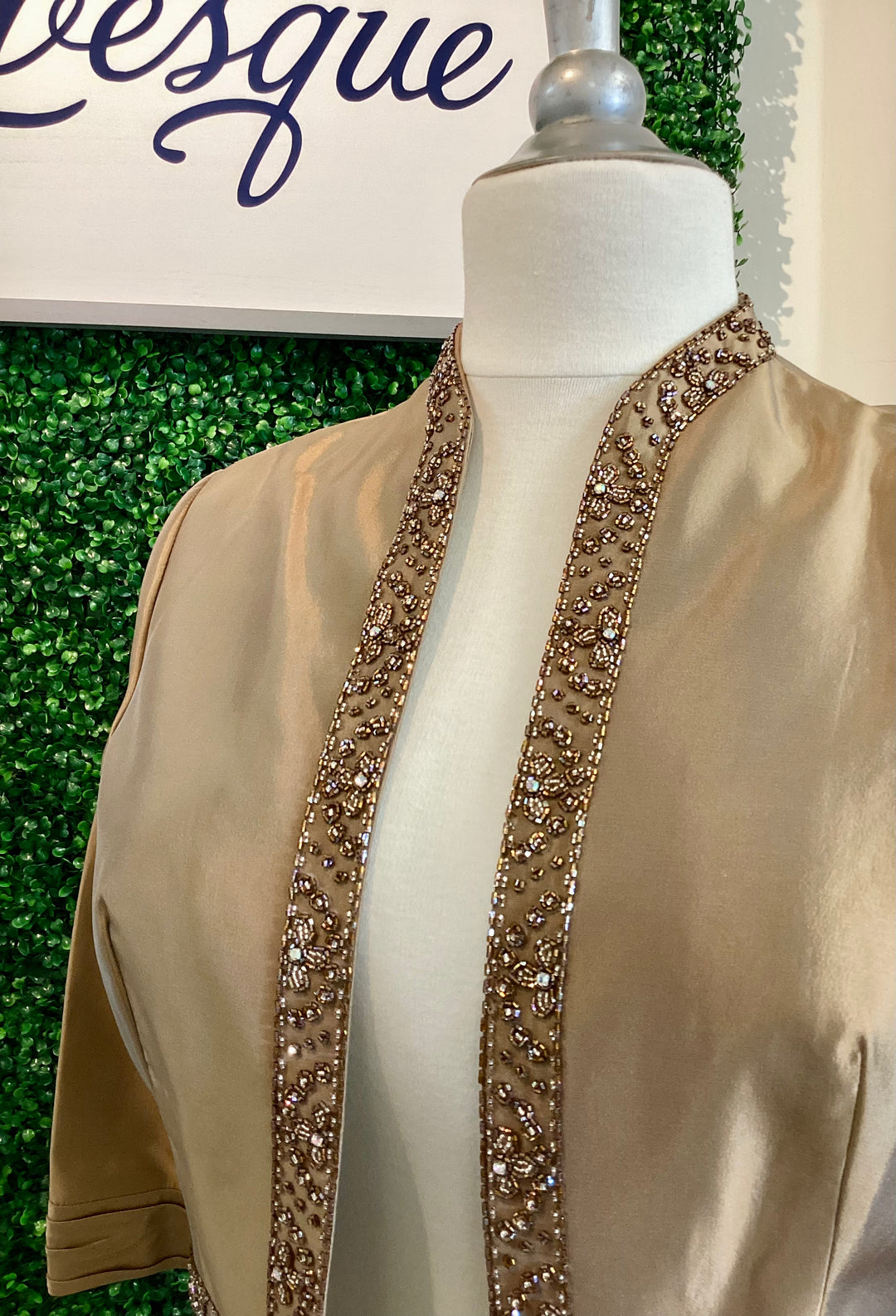 Bronze jacket with sequin trim on a mannequin against a green leafy background with &
