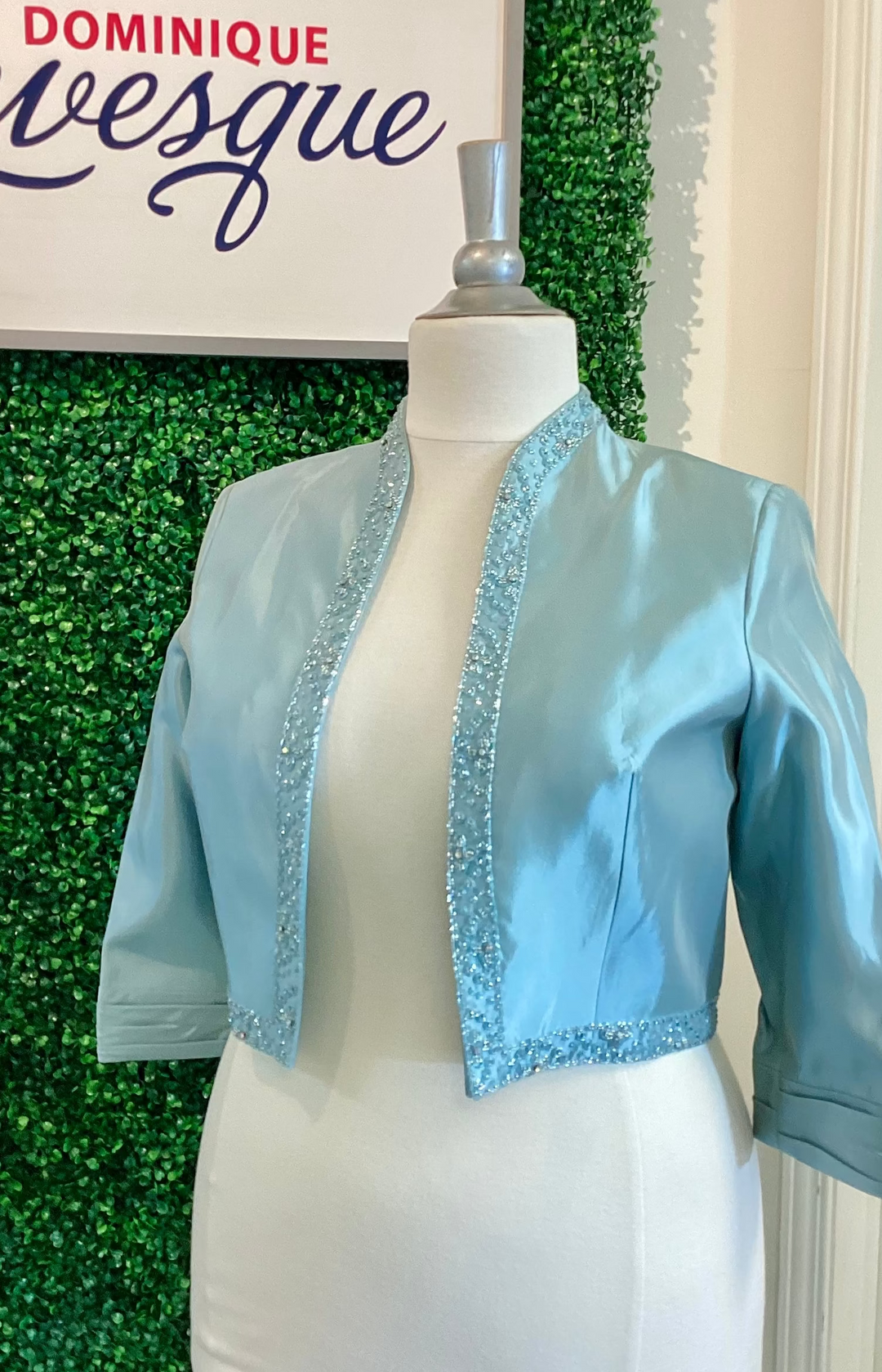 Crystal blue satin bolero jacket with sequin trim on a mannequin against a green leafy background.