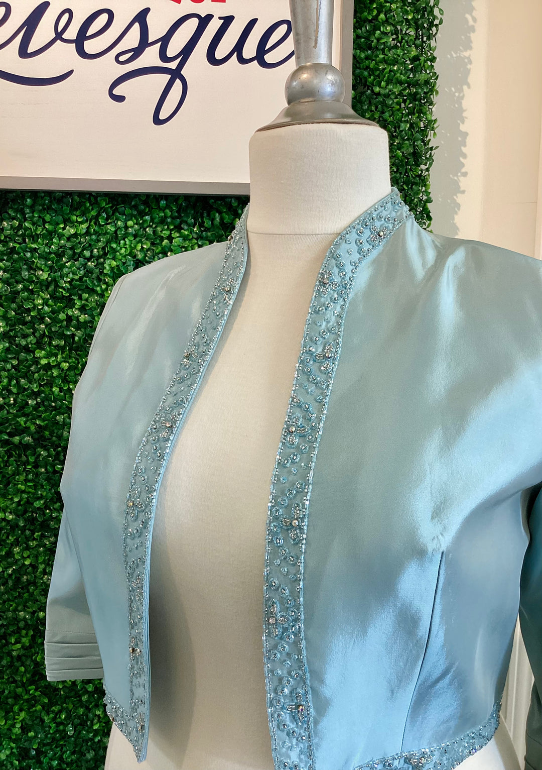 Crystal blue jacket with decorative trim on a mannequin against a green leafy background.