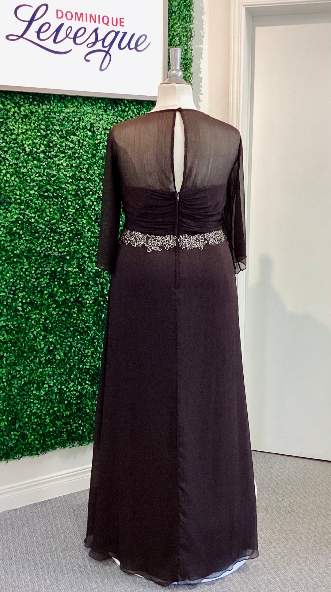 Chocolate evening gown with sheer overlay on a mannequin against a green leafy background.