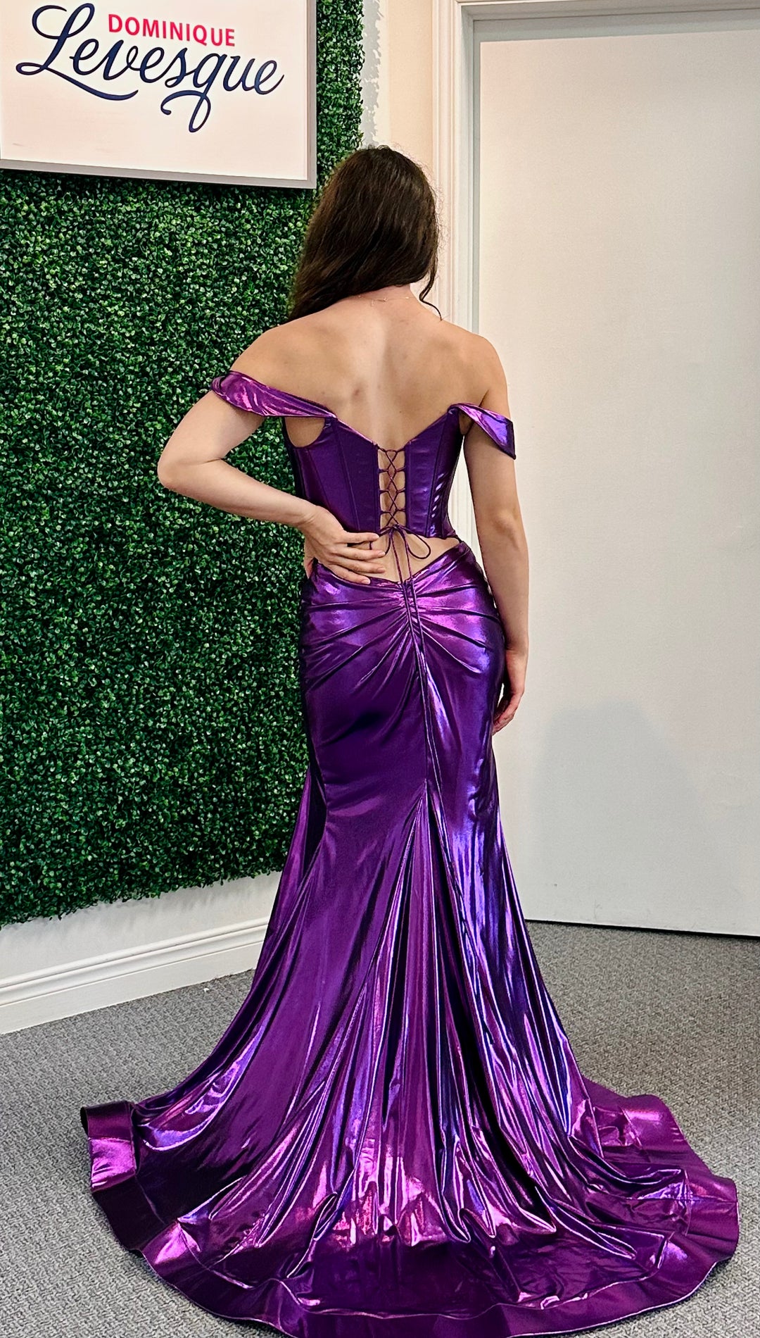 Woman in a purple off-shoulder gown with a high slit standing against a green leafy wall.