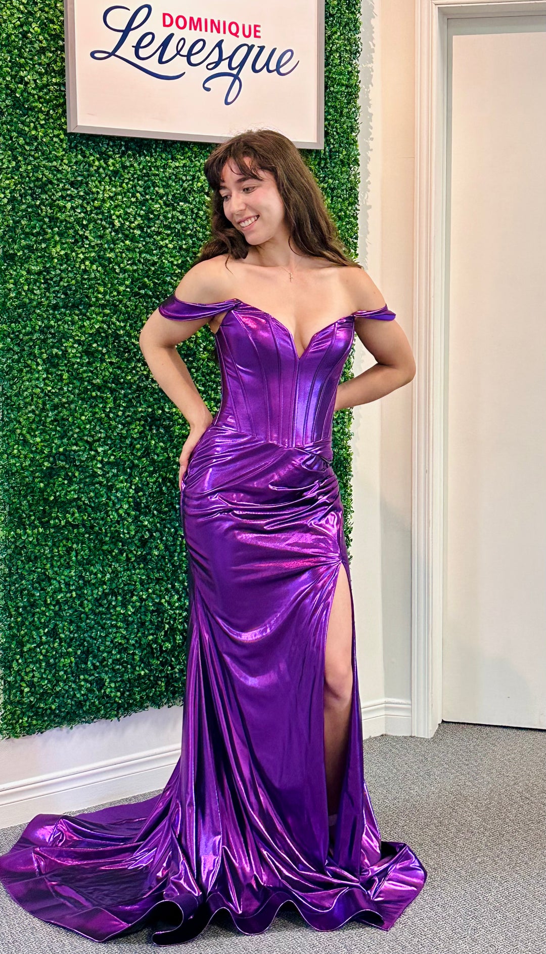 Woman in a purple off-shoulder gown with a high slit standing against a green leafy wall.