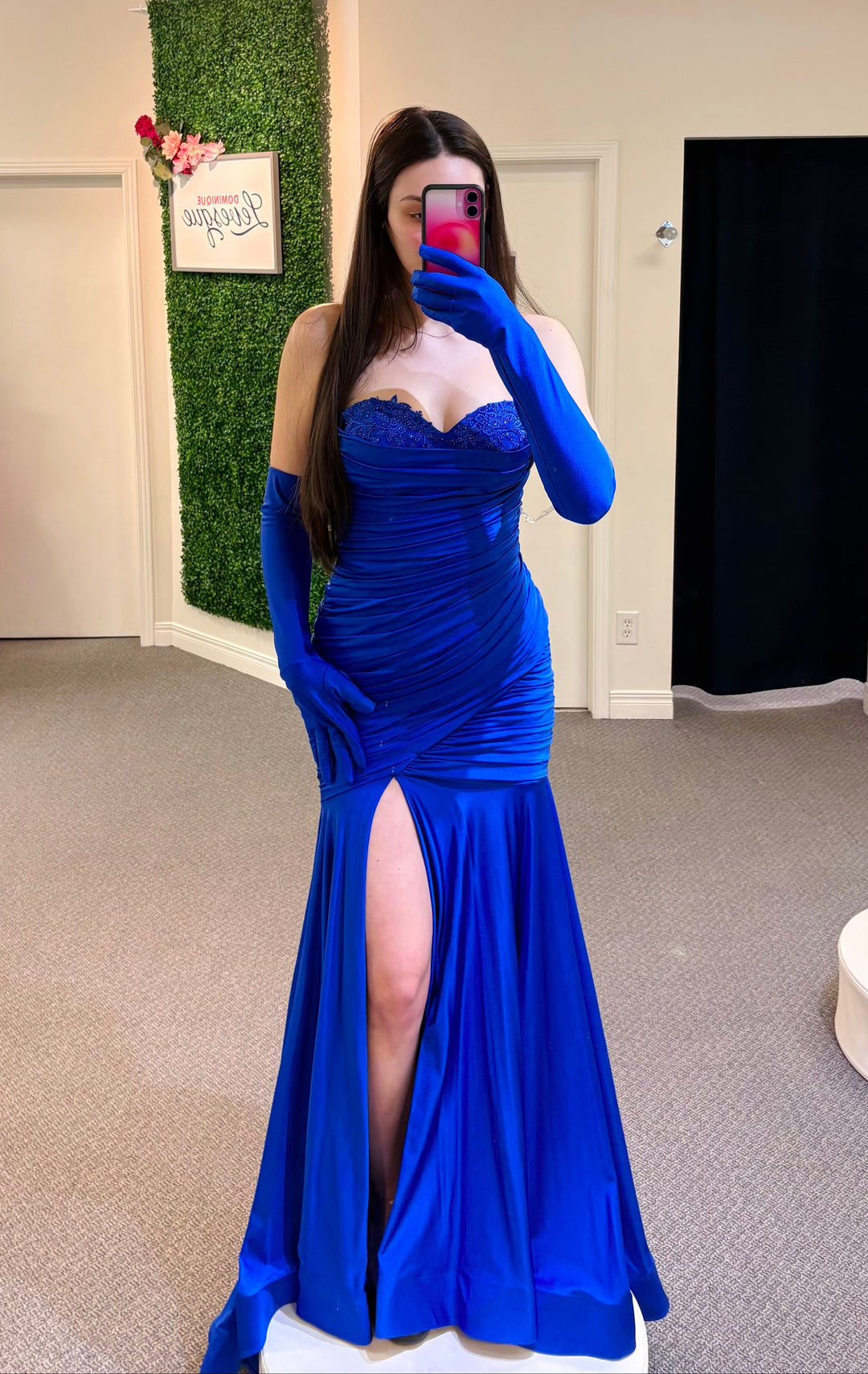 Woman wearing a blue evening gown with a high slit, standing in a fitting room.