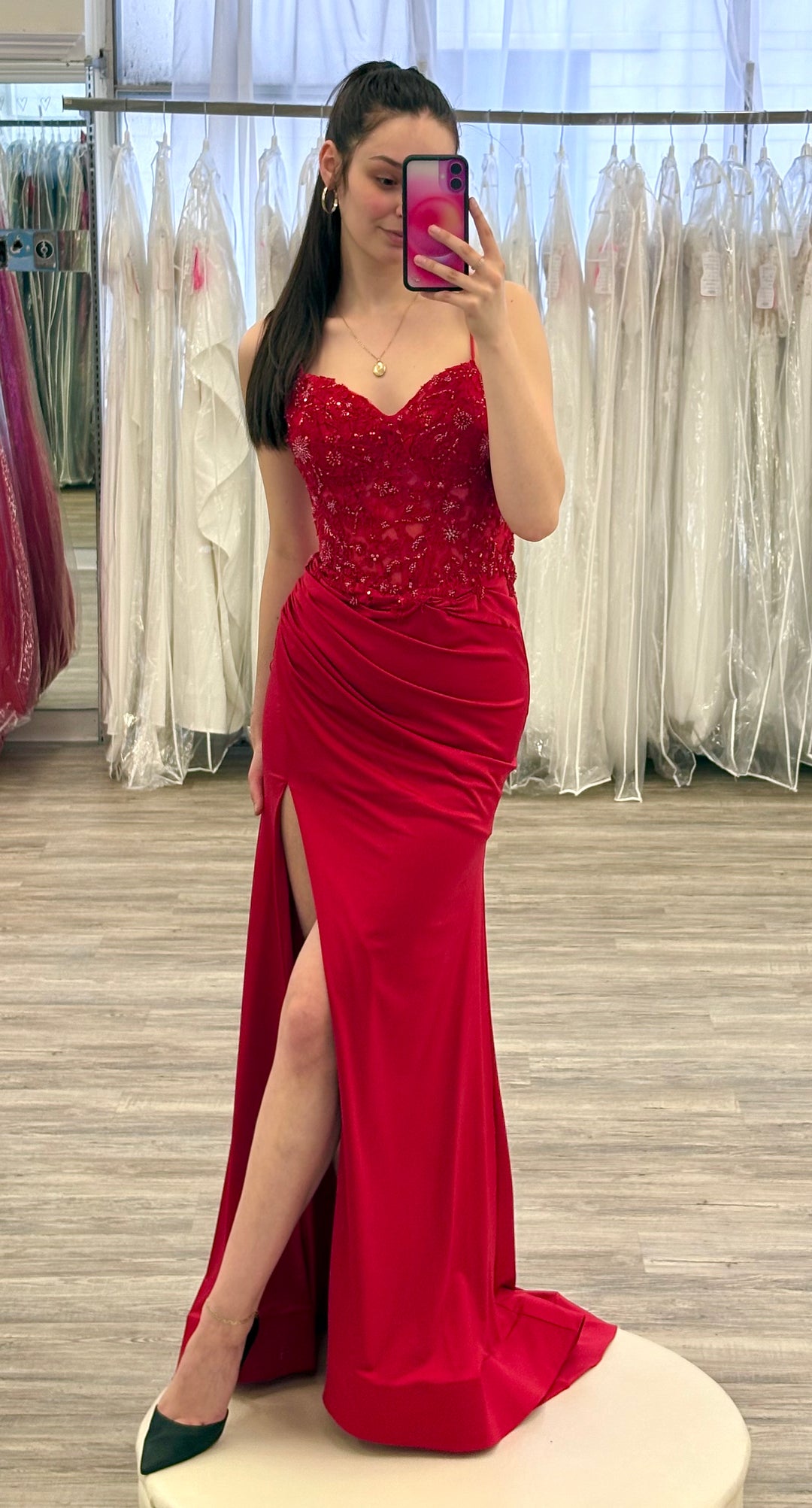 Woman in a red evening gown taking a mirror selfie in a fitting room.
