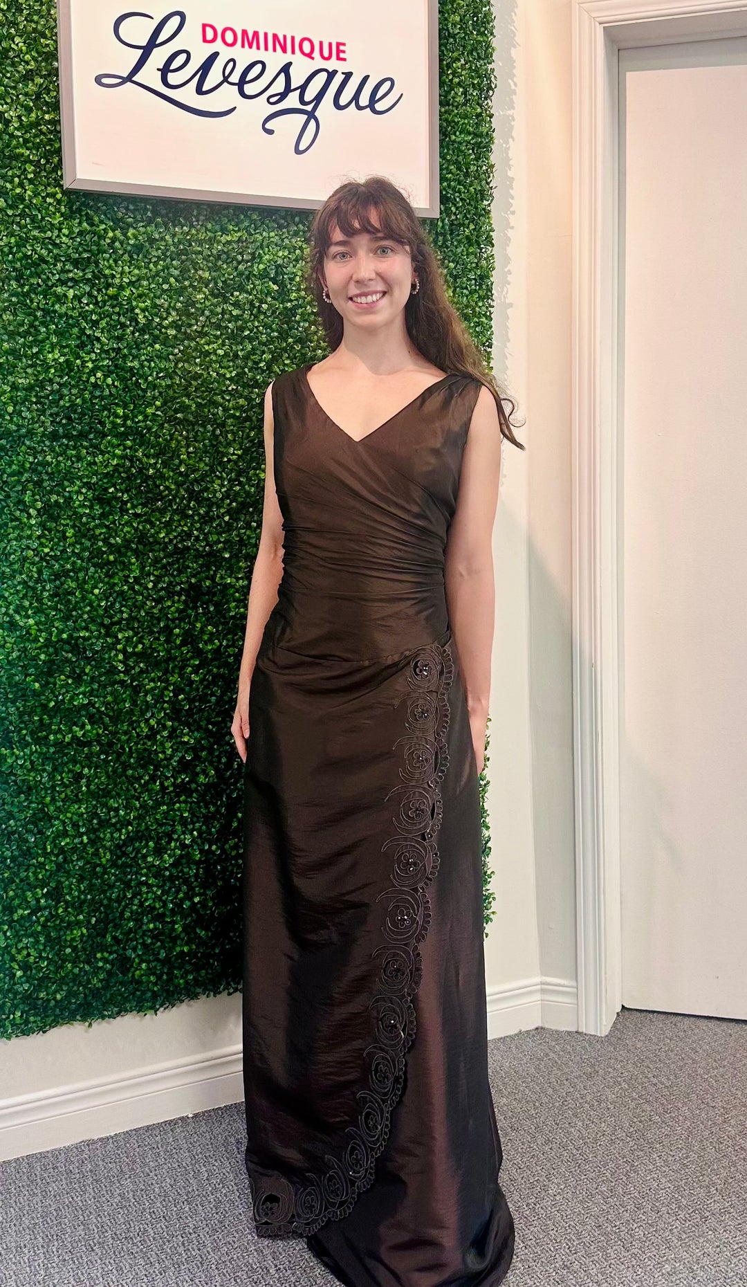 Woman in a brown evening gown standing against a green wall with a sign above saying &