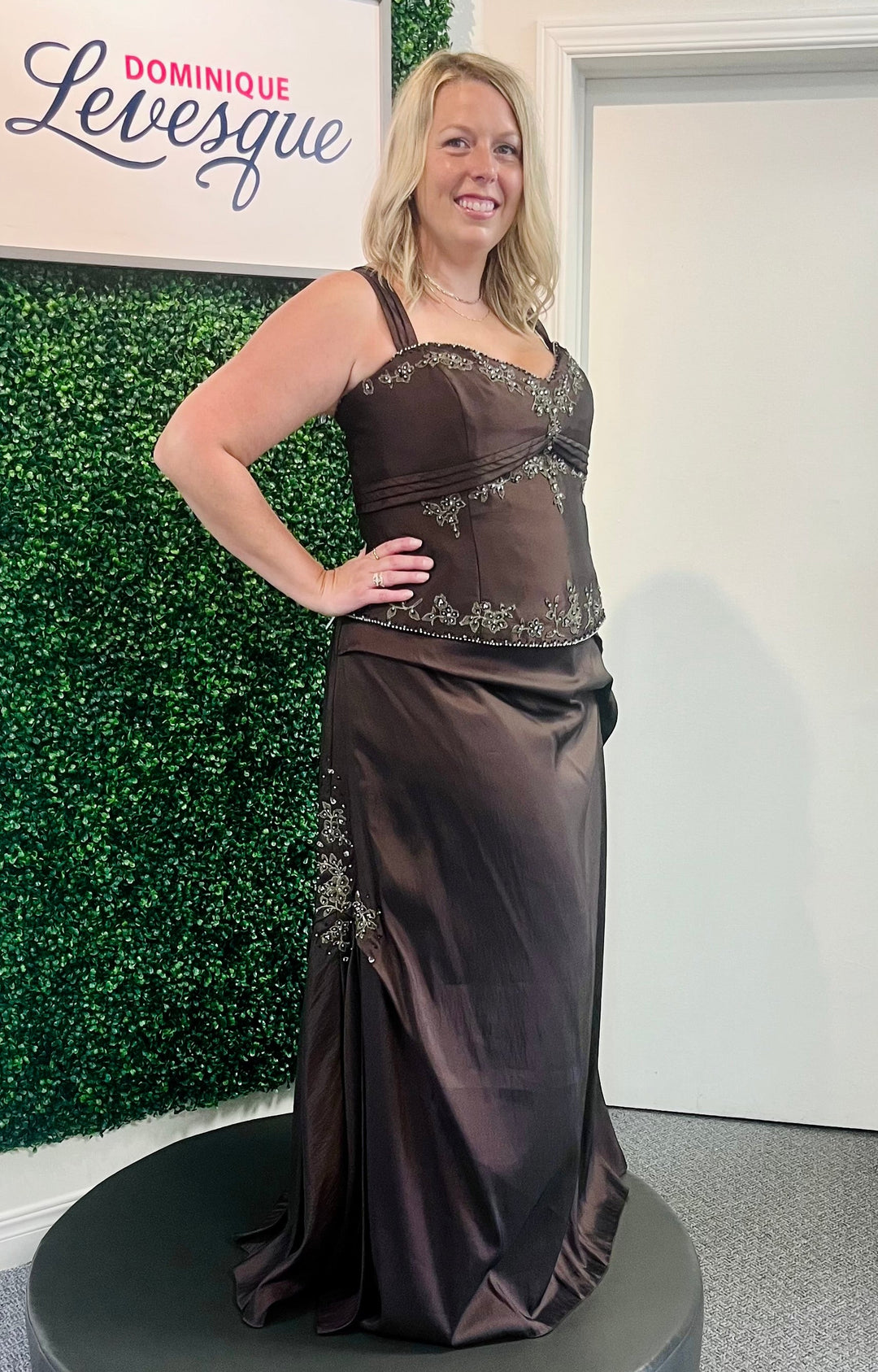 Woman wearing a brown evening gown with floral details, standing in front of a green wall and white door.