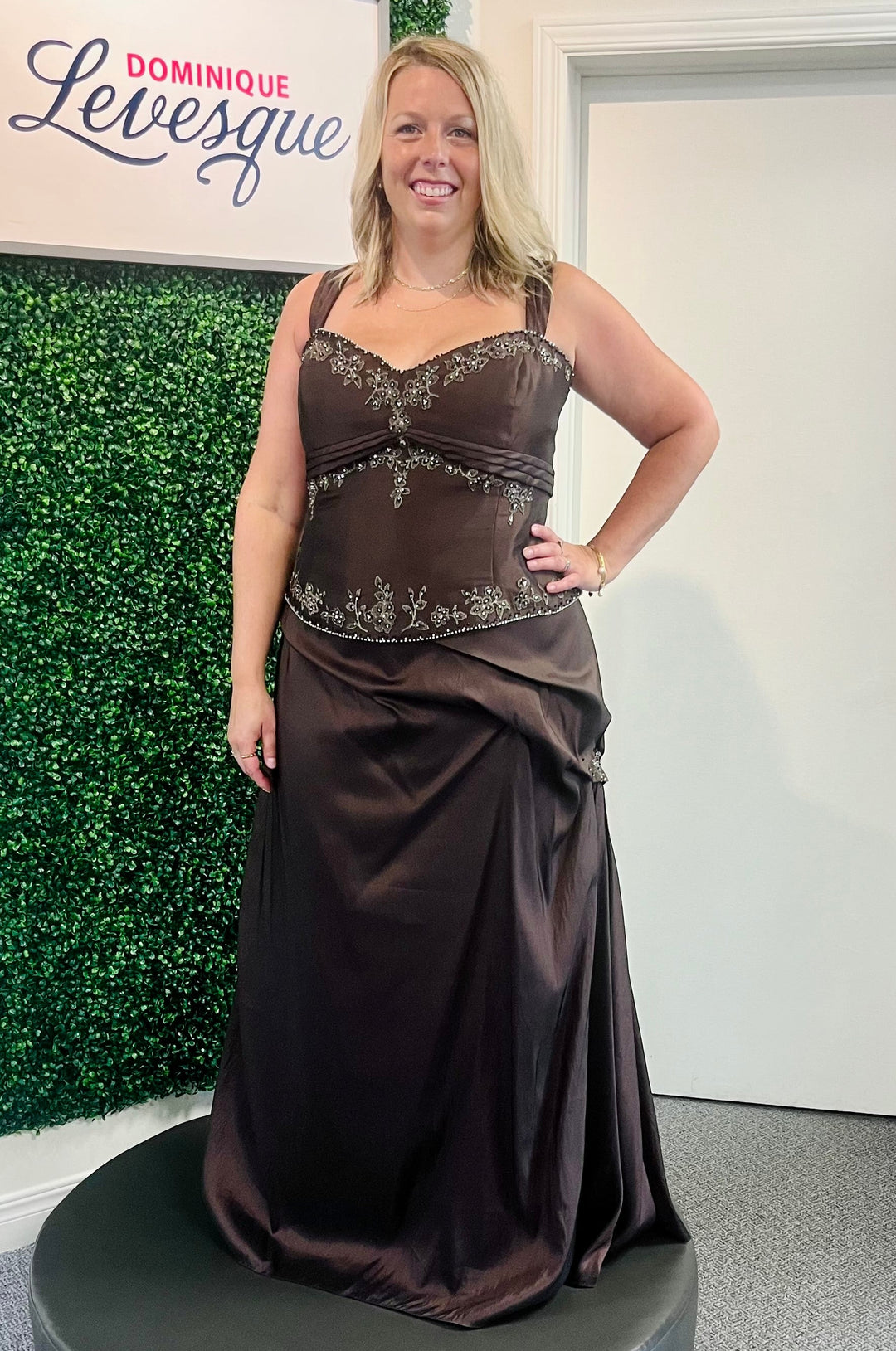 Woman wearing a brown evening gown with floral embroidery, standing against a green wall.