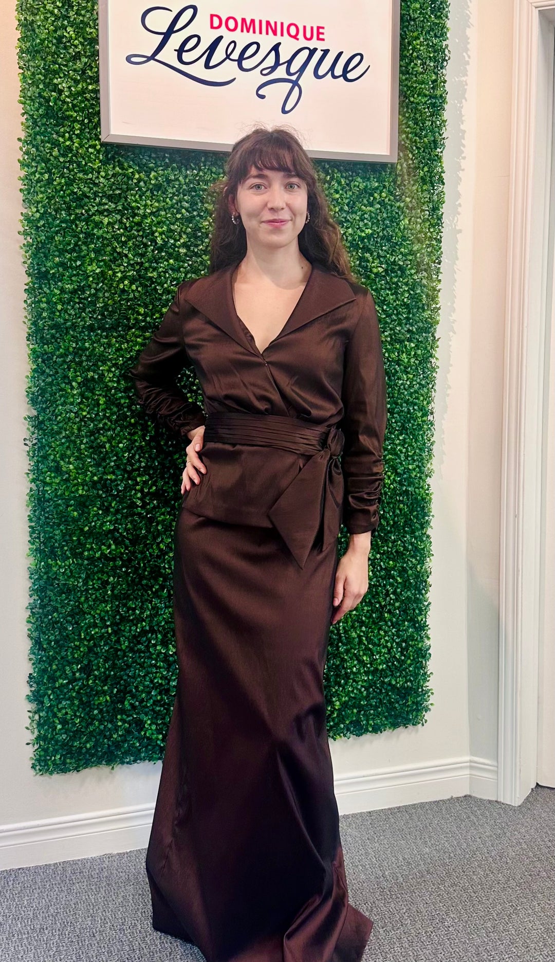 Woman in a brown dress standing in front of a green wall with a sign that reads 'Dominique Levesque'.