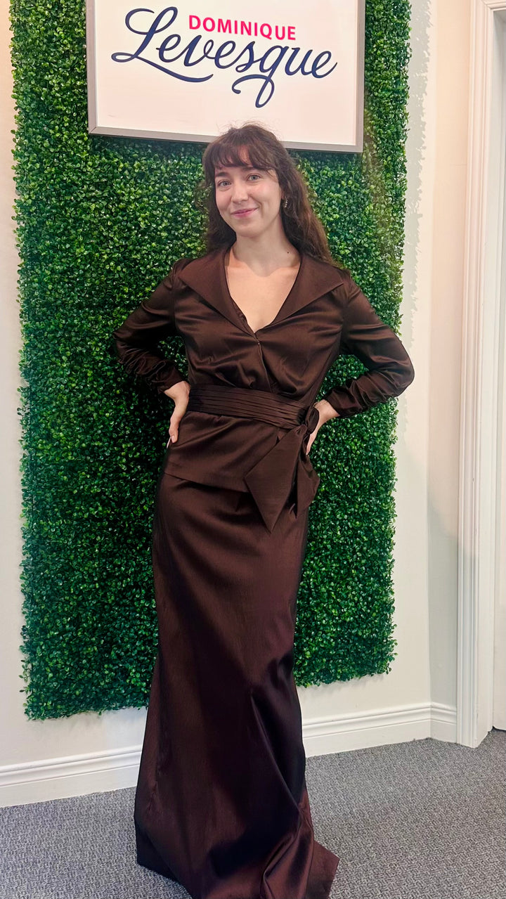 Woman in a brown dress standing in front of a green wall with a sign above saying &