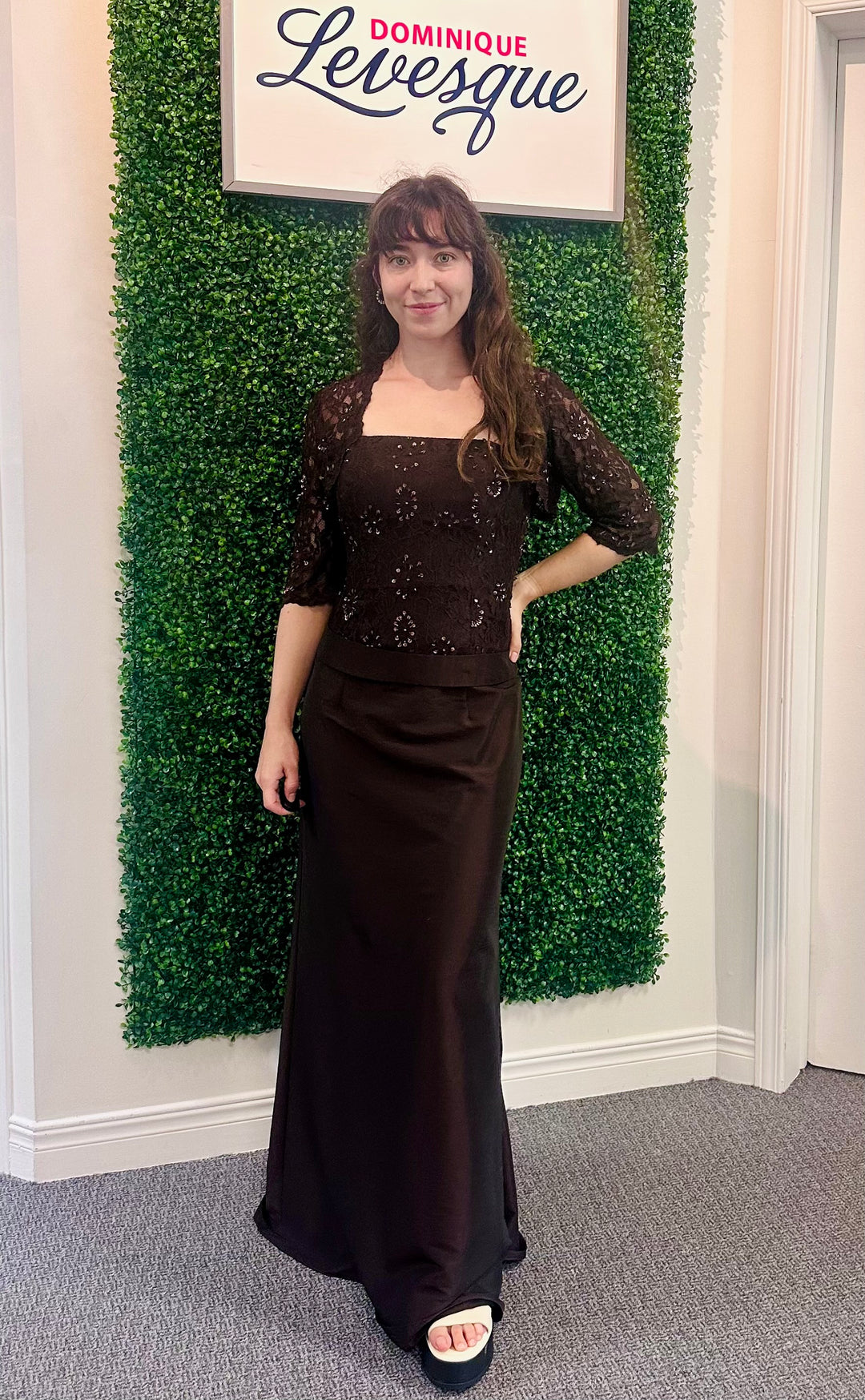 Woman in a brown dress standing in front of a green wall with a sign above it saying ''Dominique Levesque''.