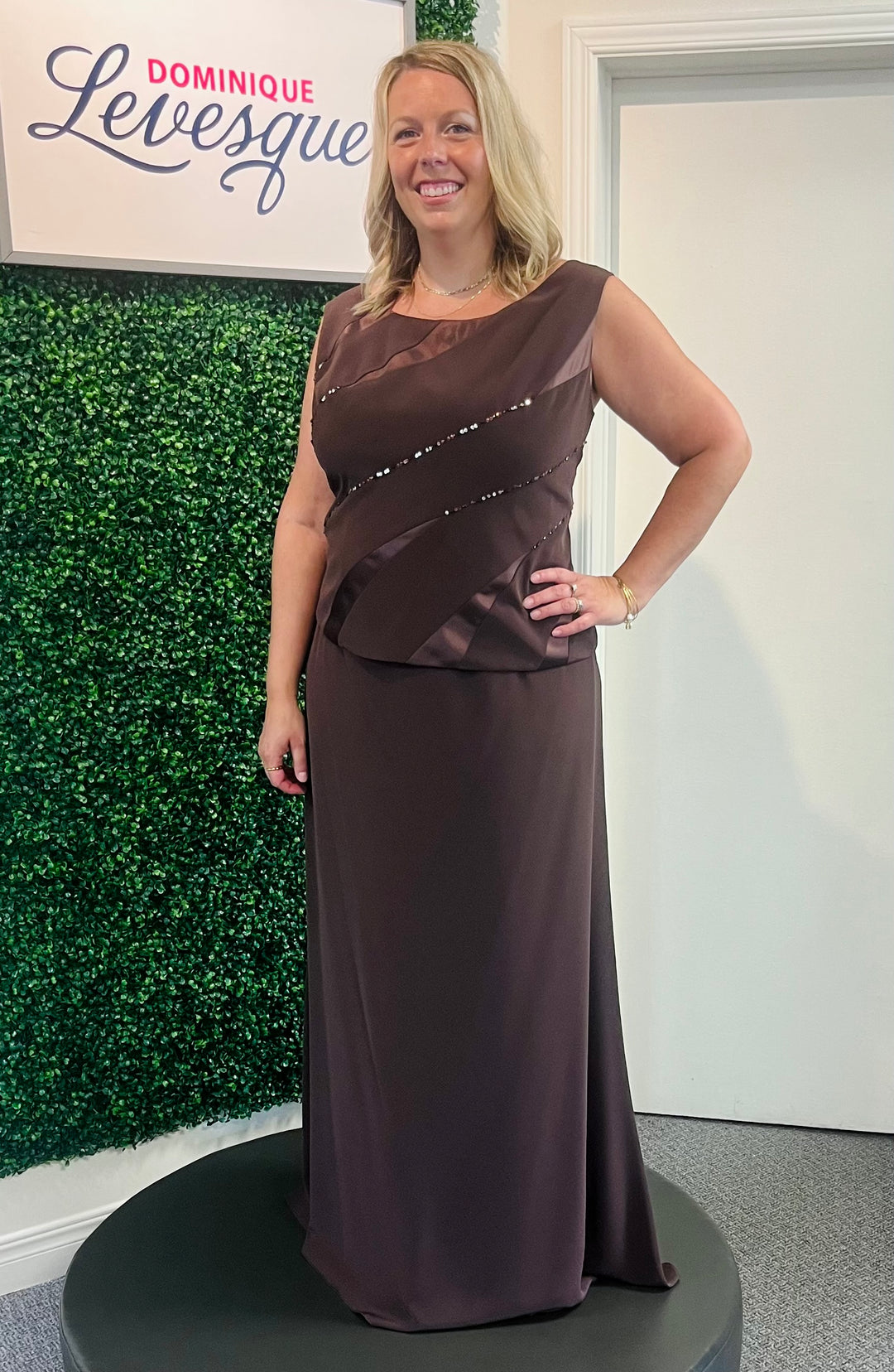 Woman in a brown evening gown standing in front of a green wall with 'Dominique Levesque' branding.
