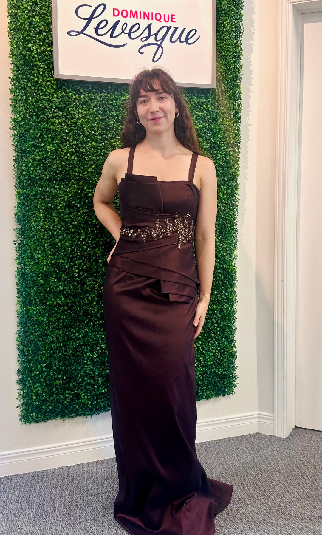Woman in a brown evening gown standing in front of a green wall with a sign above saying &