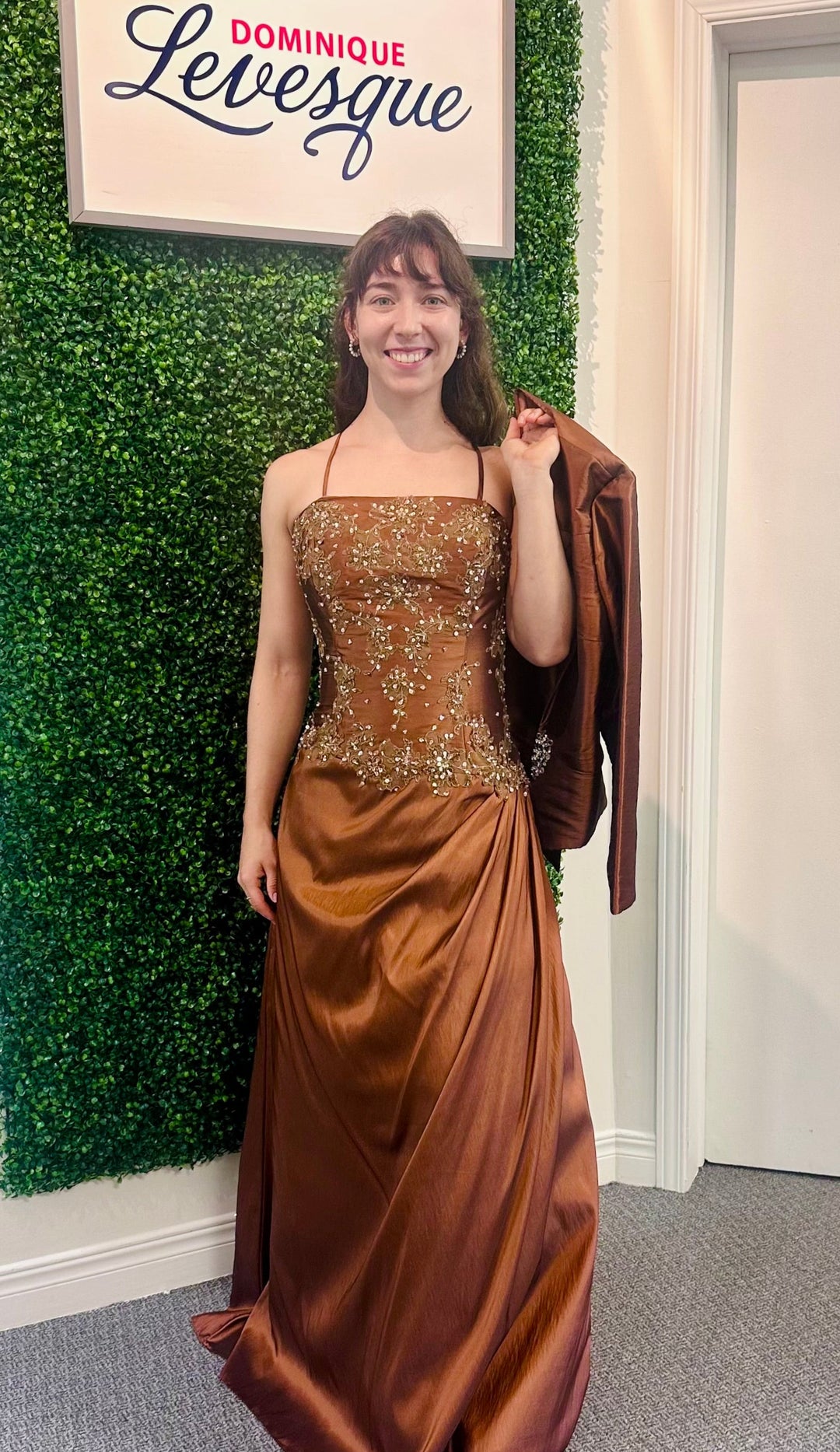 Woman in a brown evening gown standing in front of a green wall with a sign above saying 'Dominique Levesque'.