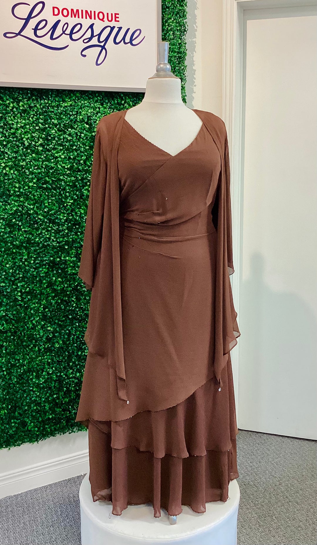 Brown dress on a mannequin against a green leafy background with 'Dominique Levesque' branding.