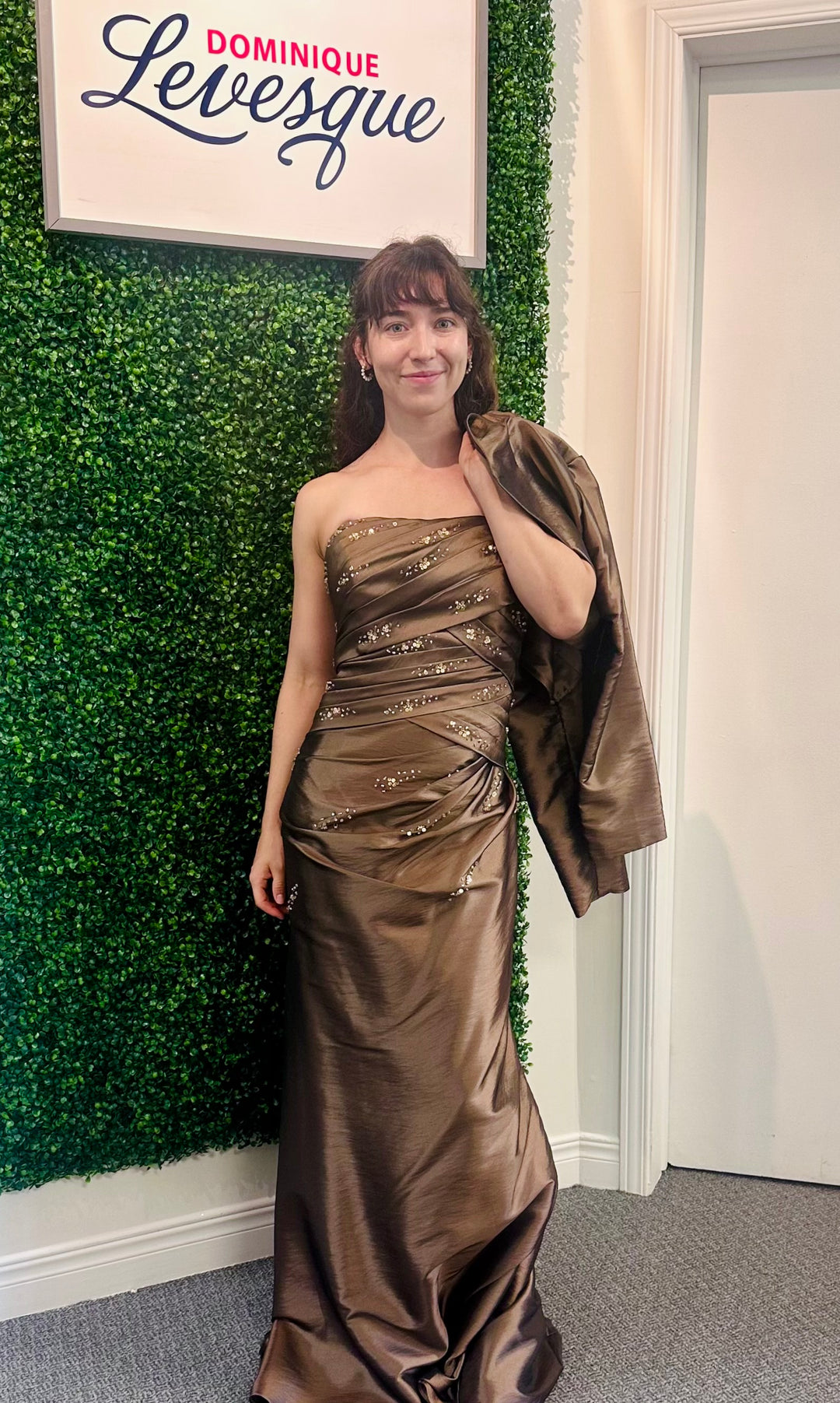 Woman in a brown strapless gown standing against a green wall with a sign above.