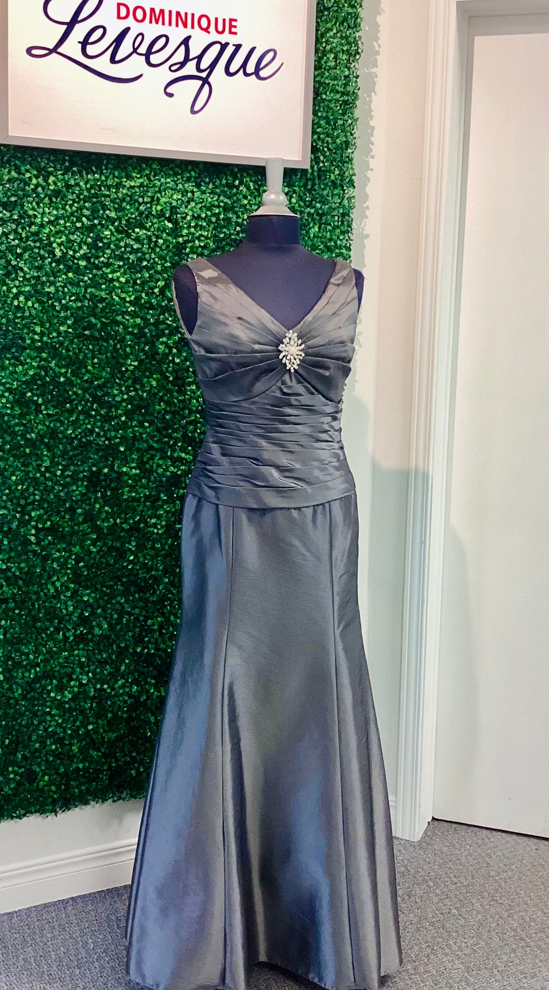 Gray evening gown on a mannequin against a green leafy background with 'Dominique Levesque' branding.