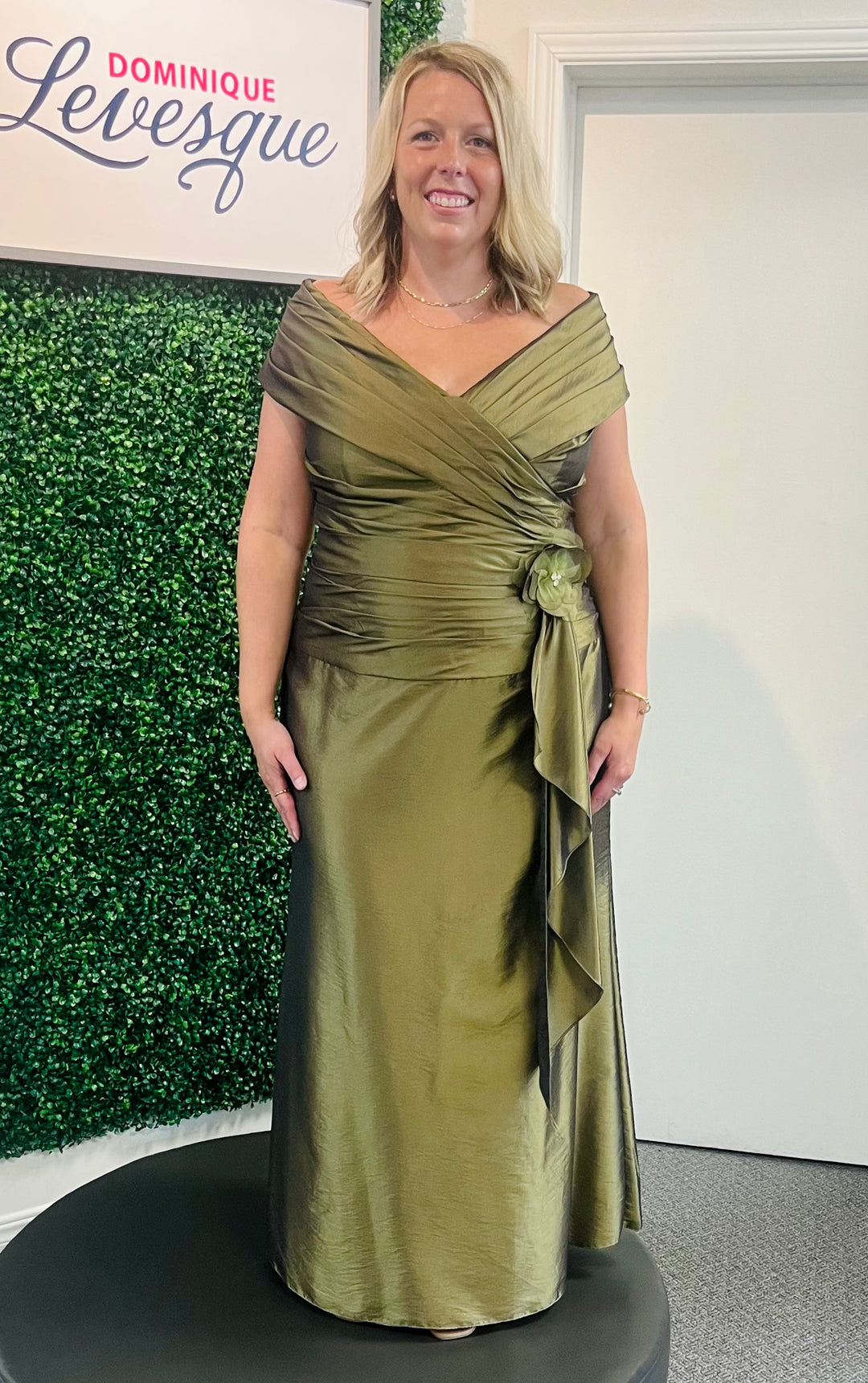 Woman in a green dress standing in front of a green wall with 'Dominique Levesque' branding.