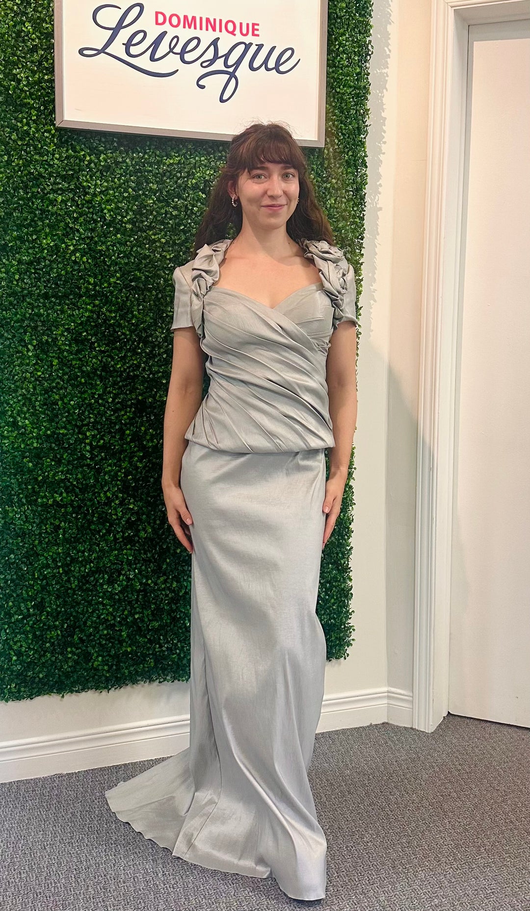 Woman in a silver dress standing against a green leafy wall with a sign above.