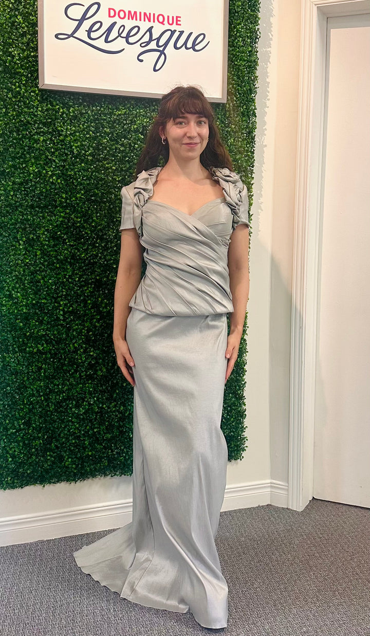 Woman in a silver dress standing against a green leafy wall with a sign above.