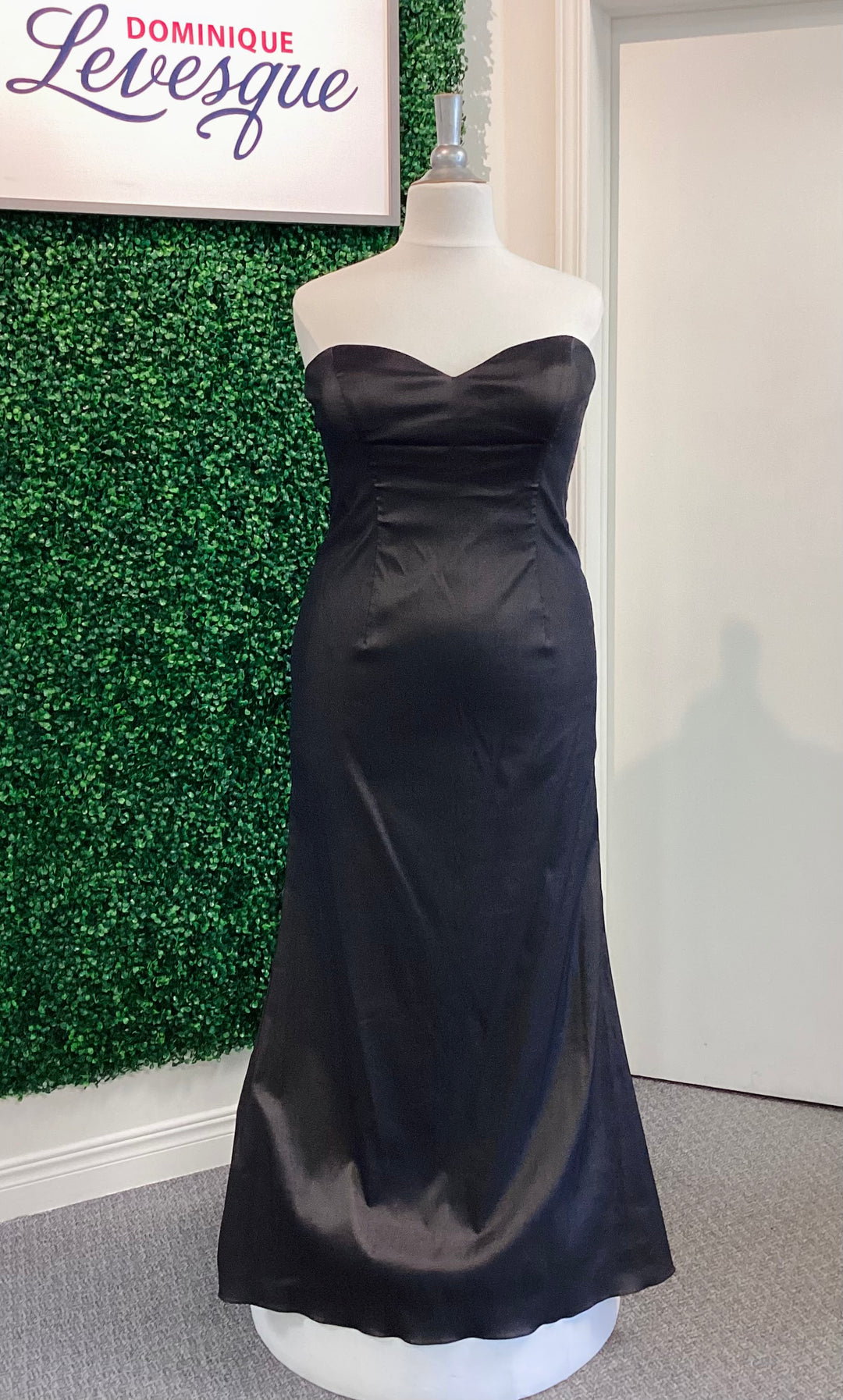 Black strapless gown on a mannequin against a green leafy background with &