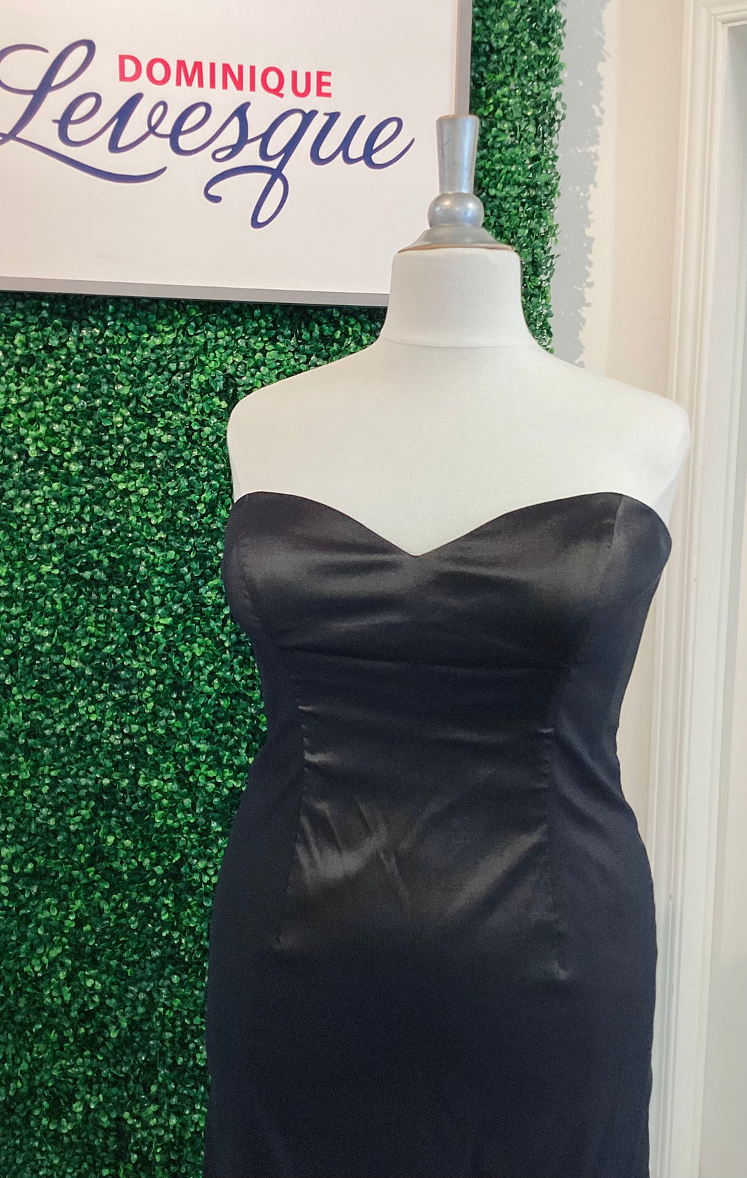Black strapless gown on a mannequin against a green leafy background with &