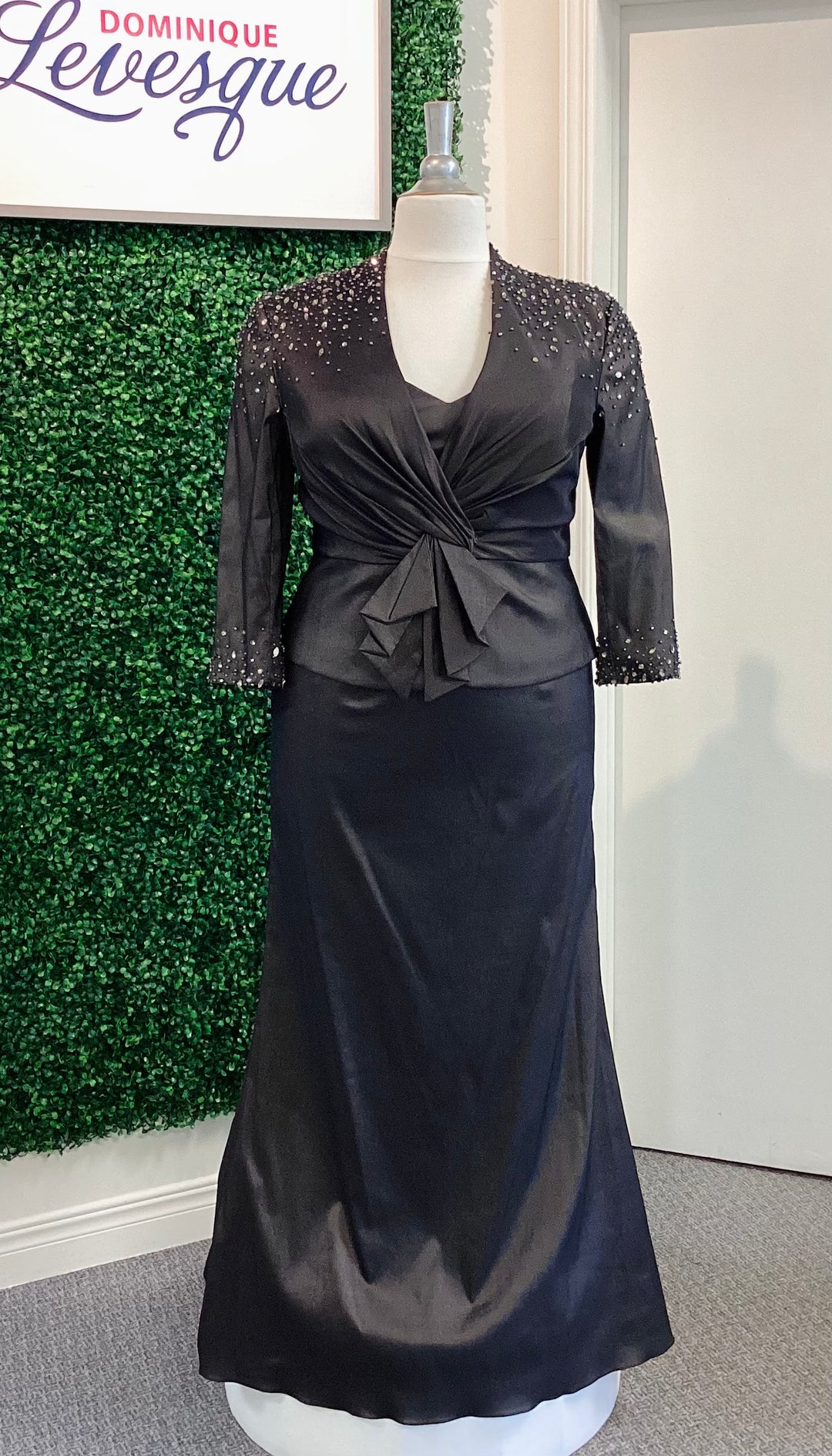 Black strapless gown with vest on a mannequin against a green leafy background with 'Dominique Levesque' branding.