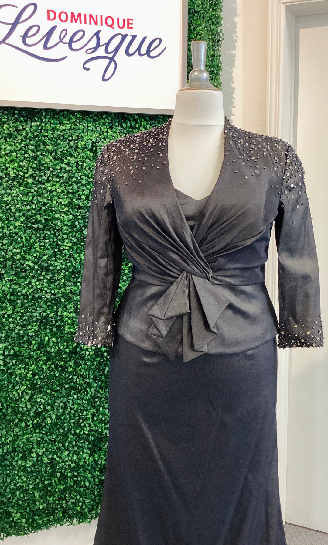 Black strapless gown with vest on a mannequin against a green leafy background with &