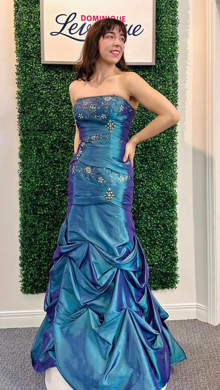 Woman in a blue and purple strapless gown posing in front of a green wall with a sign.