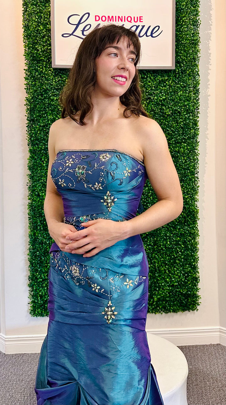 Woman in a blue and purple strapless gown posing in front of a green wall with a sign.