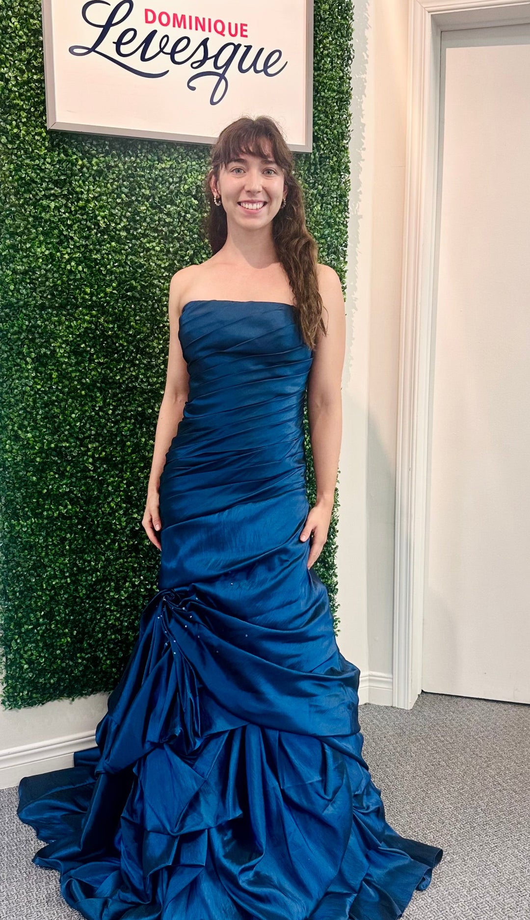 Woman in a blue strapless gown standing against a green leafy wall with 'Dominique Levesque' sign.