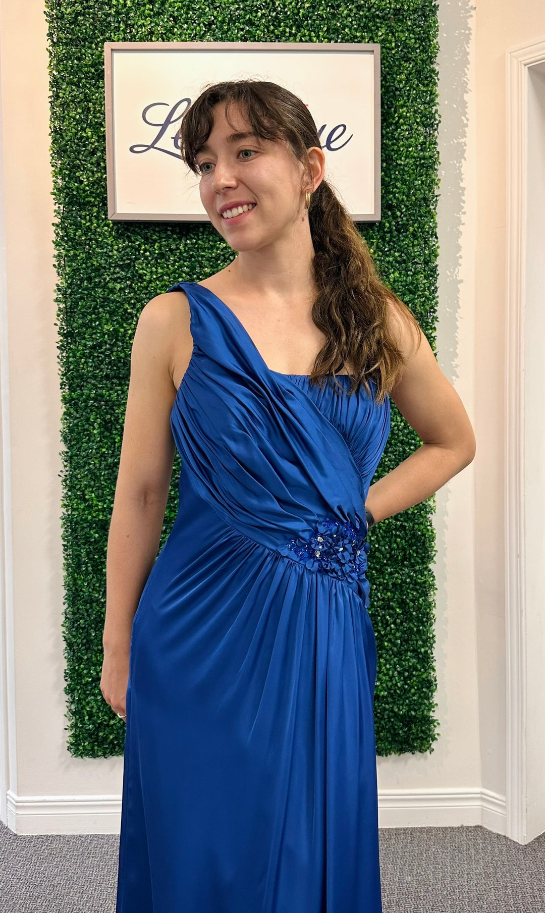Woman in a blue dress standing in front of a green leafy wall with a framed picture.
