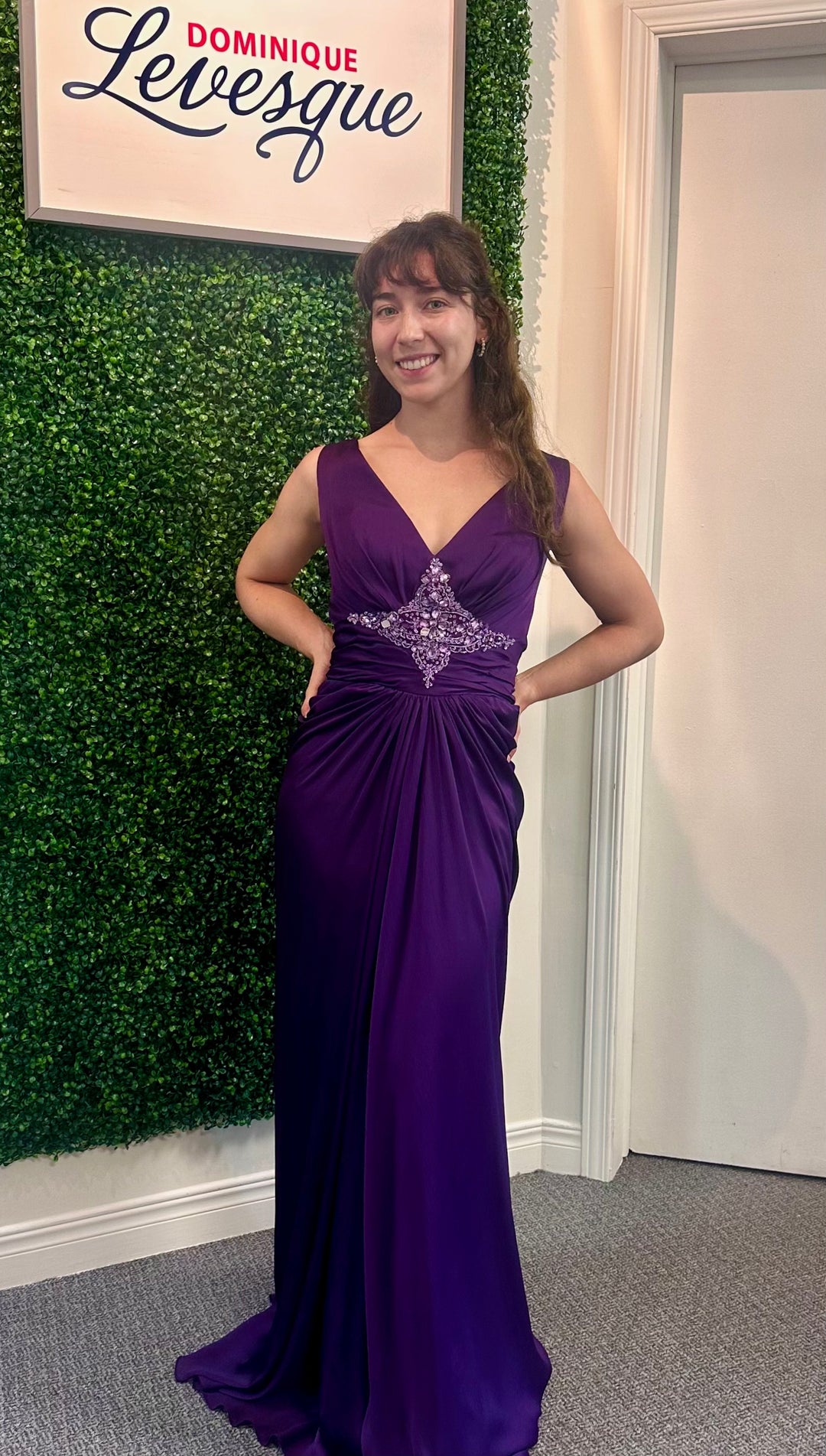 Woman in a purple evening gown standing in front of a green wall with a sign that reads 'Dominique Levesque'.