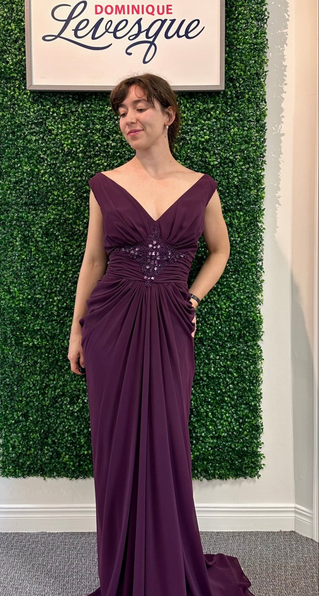 Woman in a plum evening gown standing against a green leafy background with a sign above.