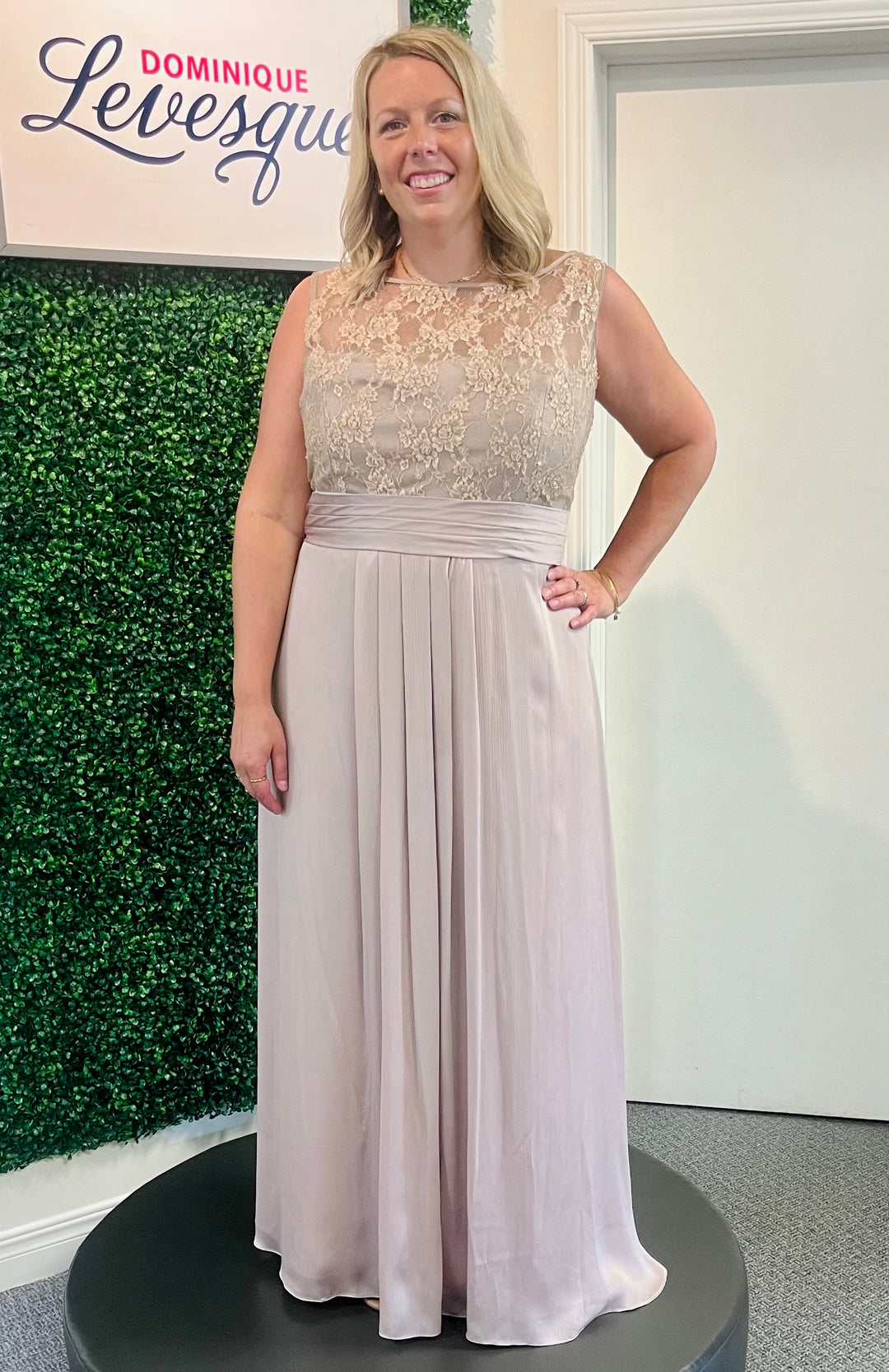 Woman wearing a long beige pink elegant dress with a green foliage background and 'Dominique Levesque' branding.