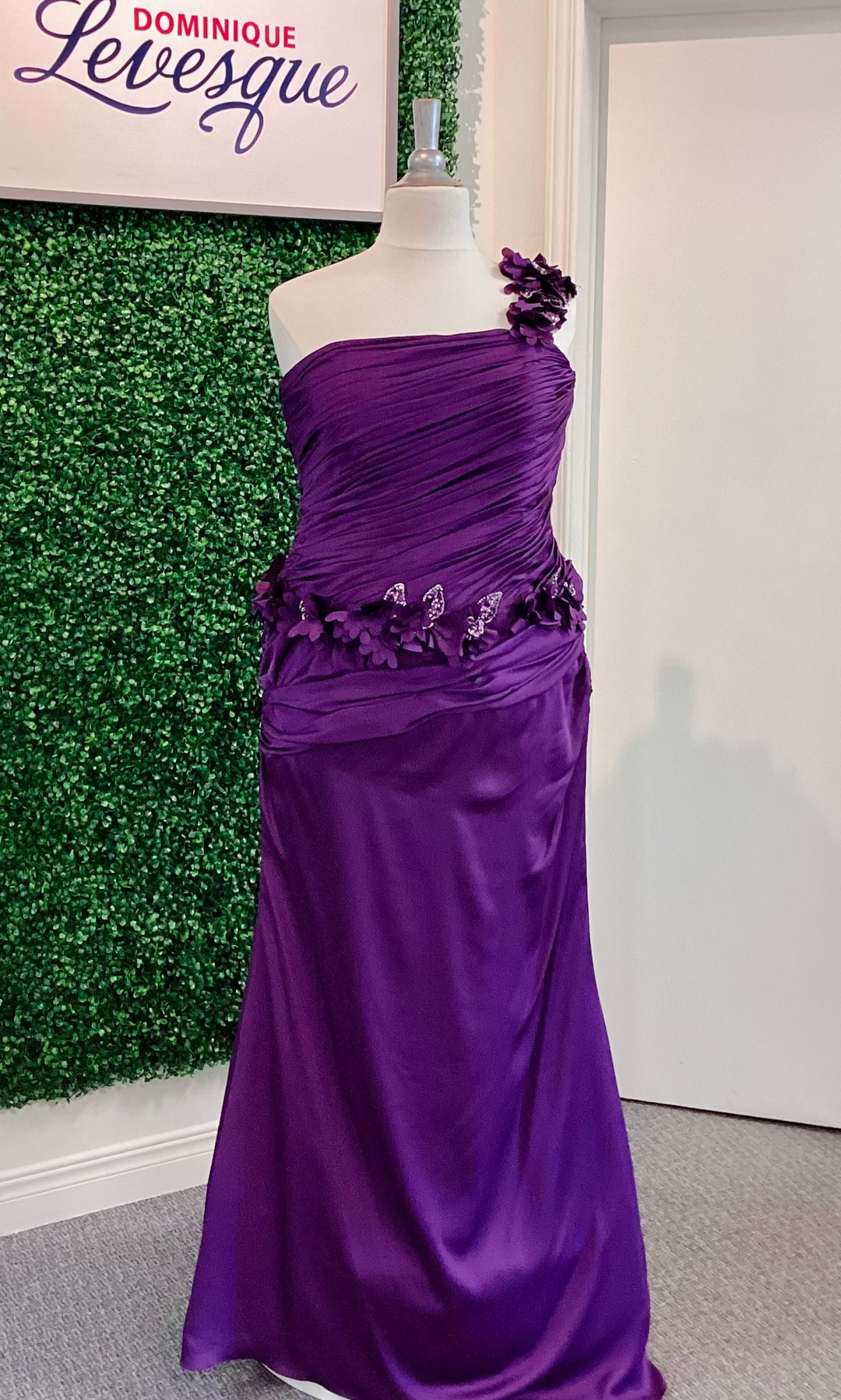 Purple evening gown on a mannequin against a green leafy background with 'Dominique Levesque' branding.