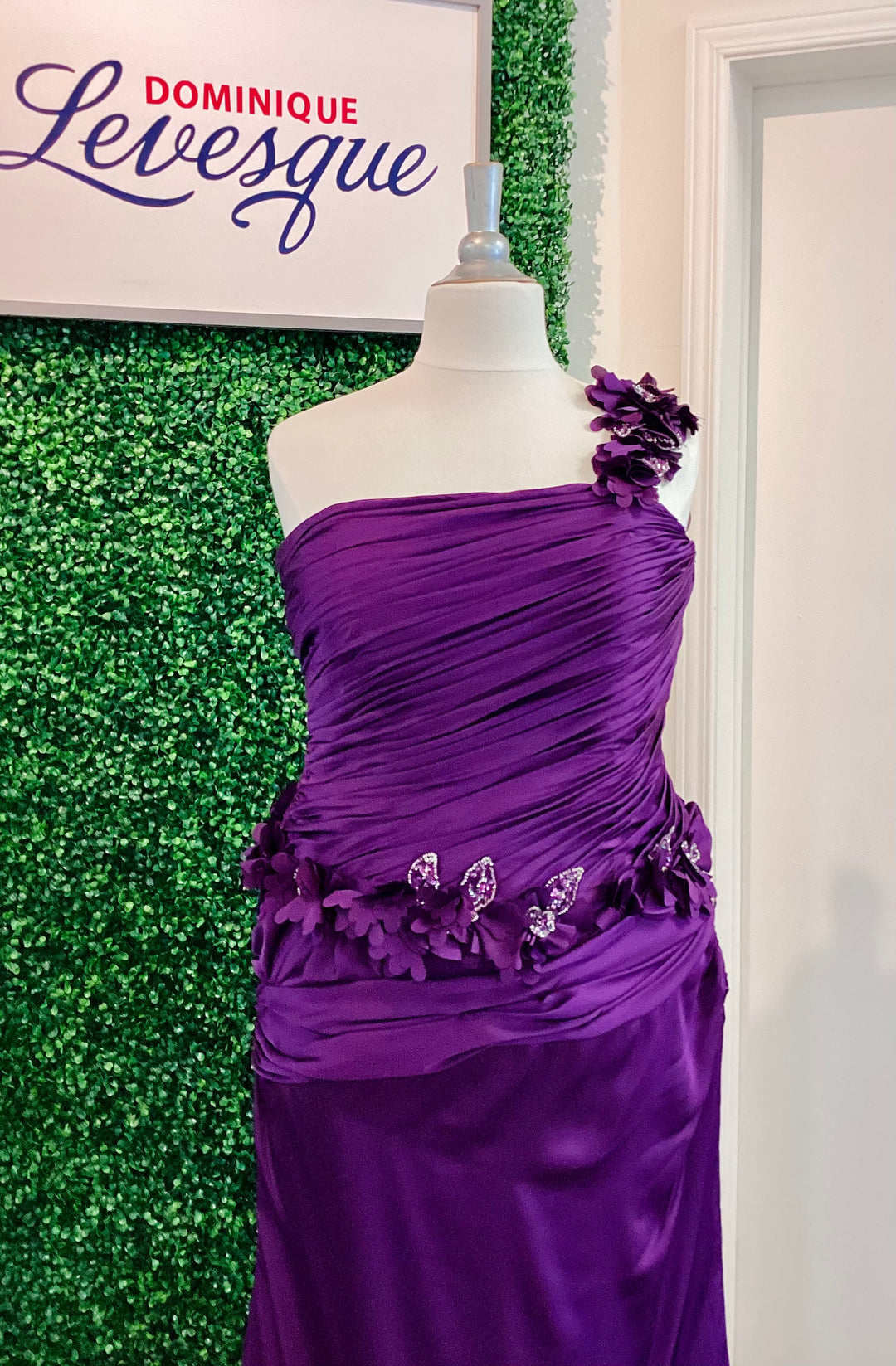 Purple one-shoulder dress on a mannequin against a green textured wall with &