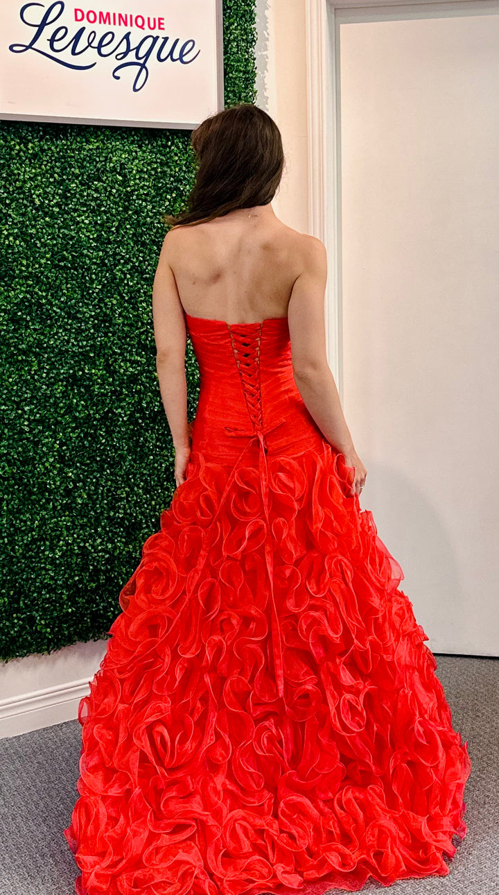 Woman wearing a red strapless gown with ruffled details, standing against a wall.