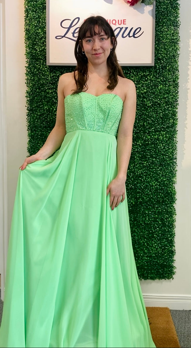Woman in a lime strapless gown standing against a green leafy background with a sign above.